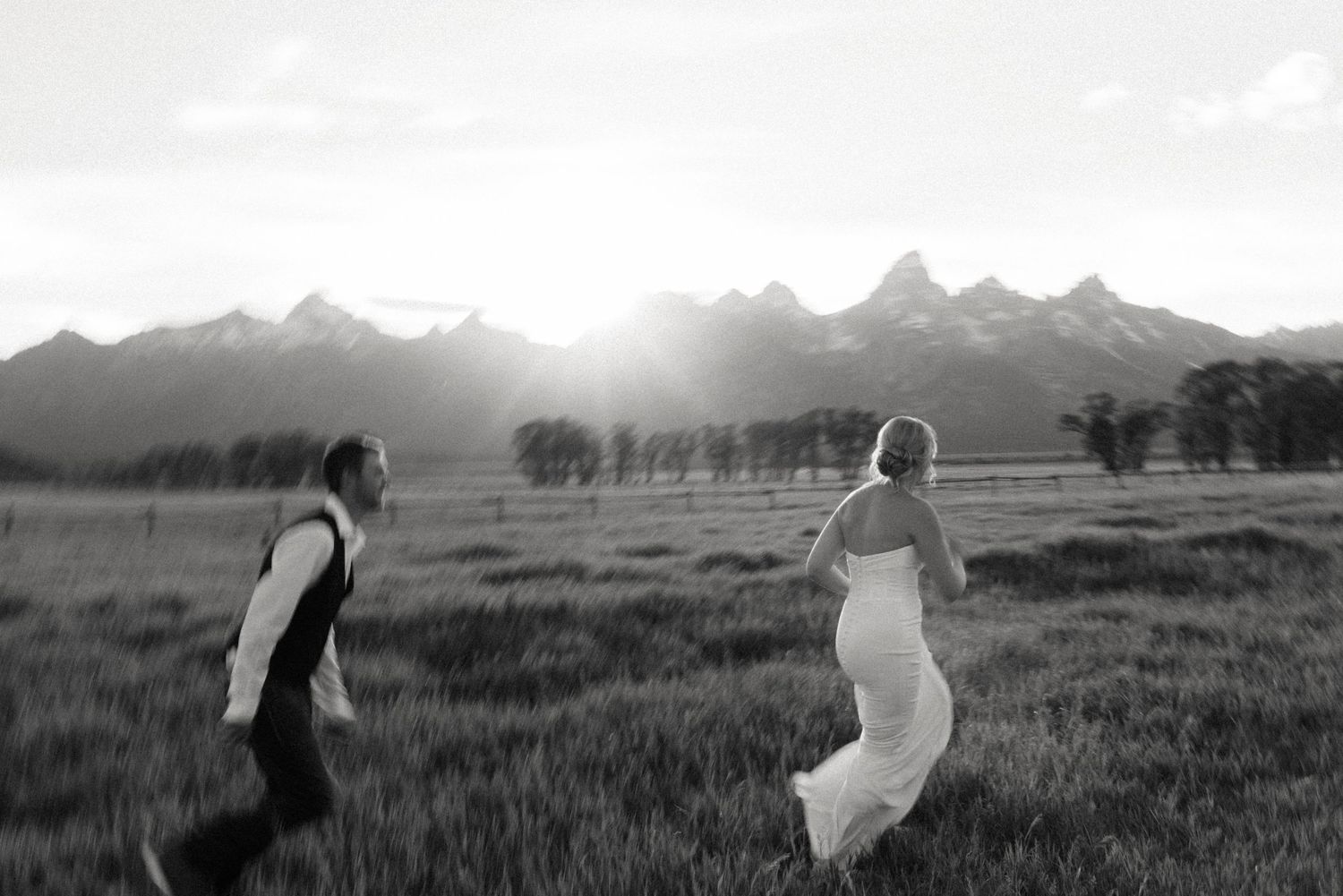Black and white photo of bride and groom running joyfully through meadow with Grand Teton mountains.
