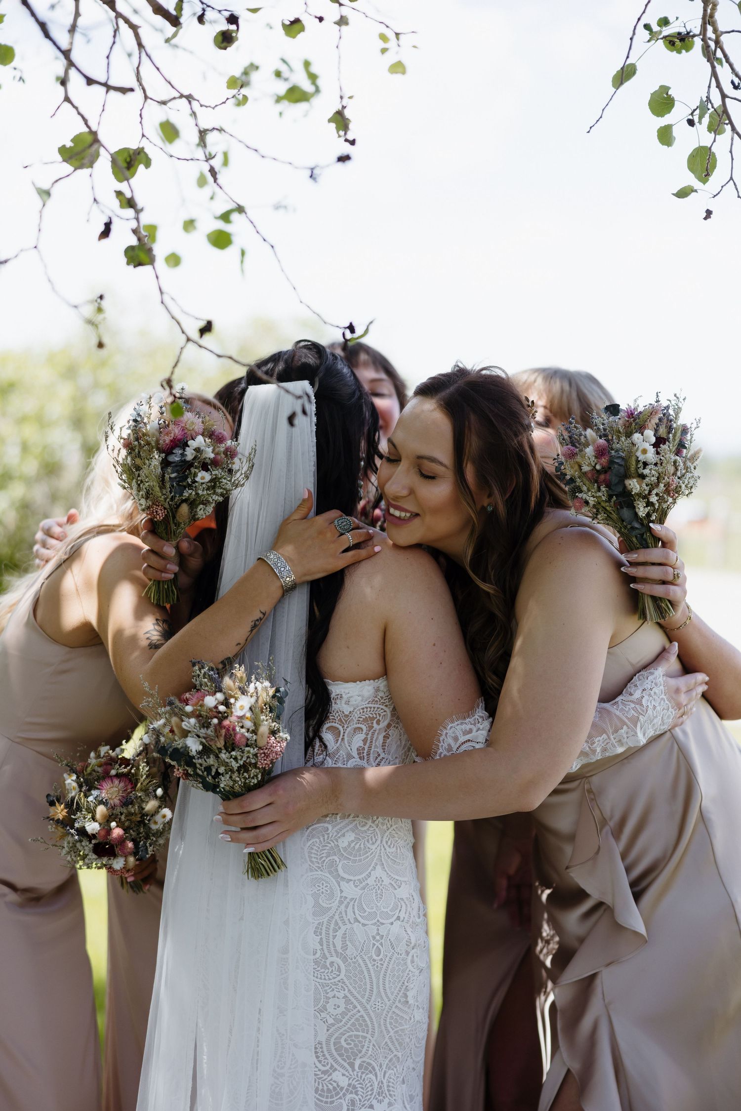 Bride in lace wedding dress shares a joyful group hug with bridesmaids holding wildflower bouquets.