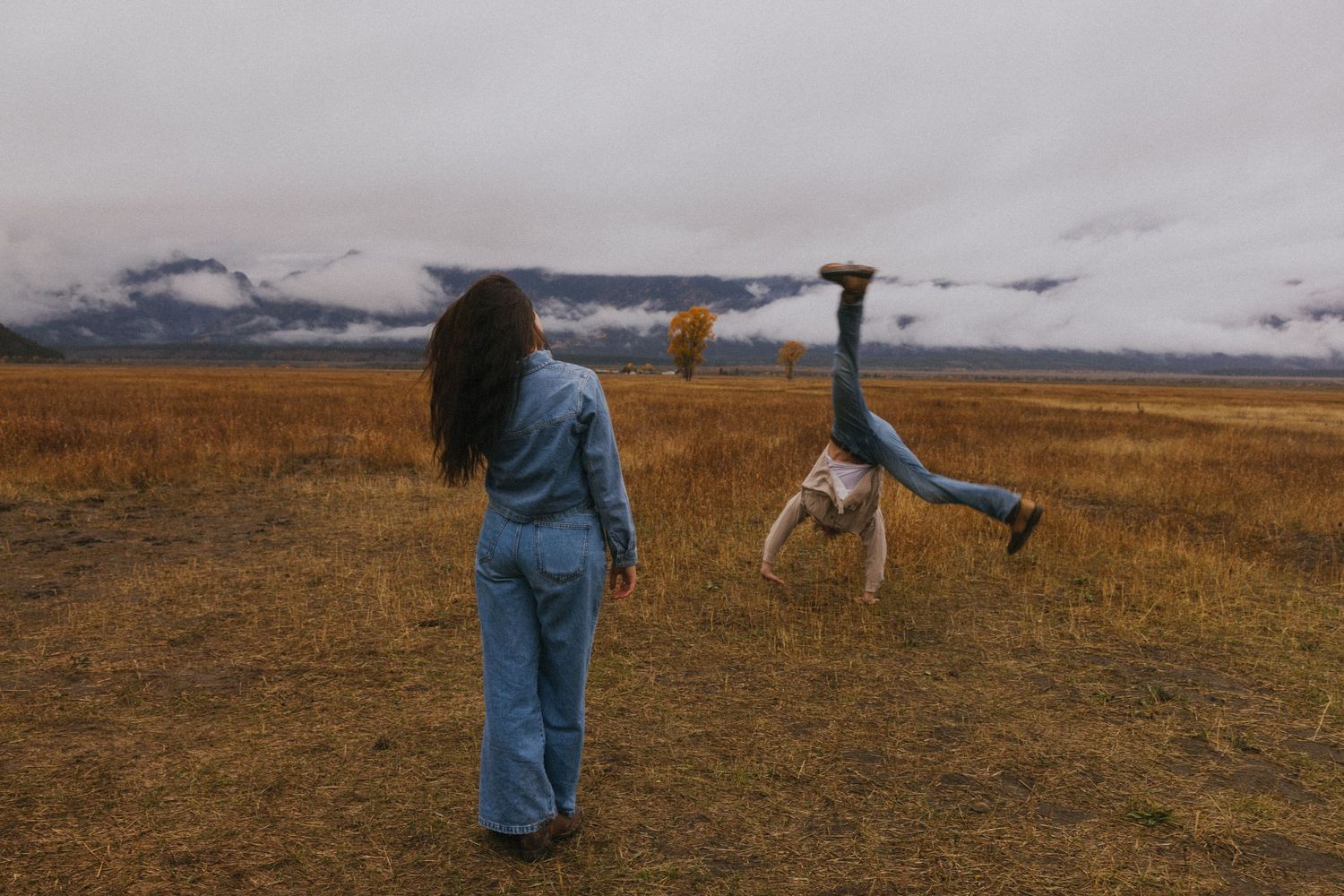 Woman watches as man does a cartwheel in a golden field with dramatic mountain backdrop.