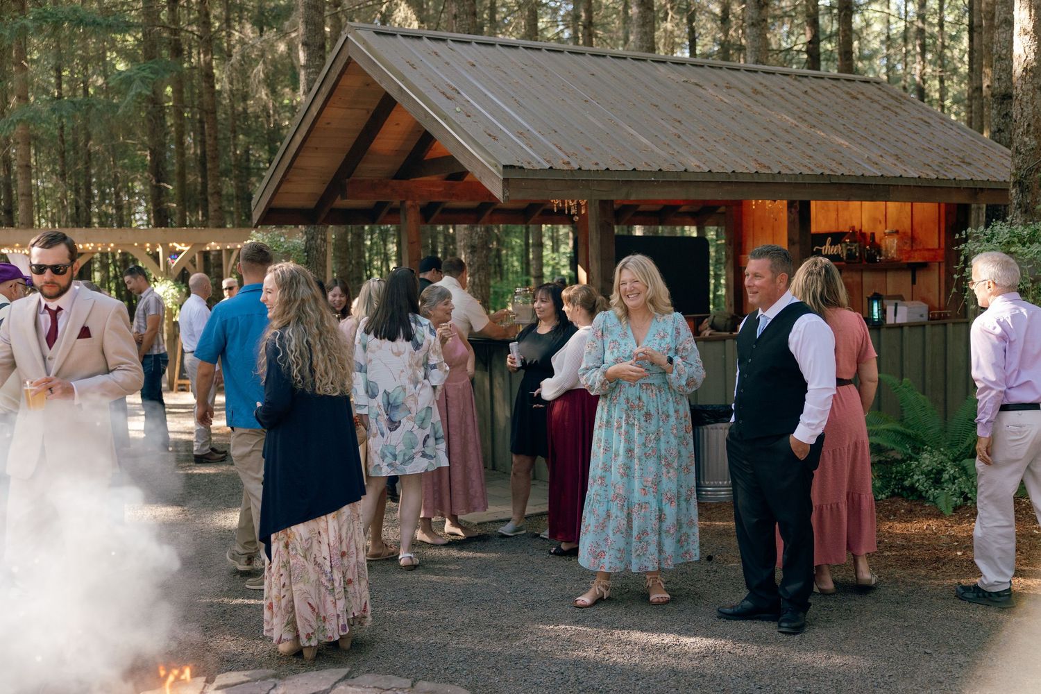 Wedding guests socialize near a rustic wooden bar pavilion surrounded by tall pine trees at an outdoor reception.