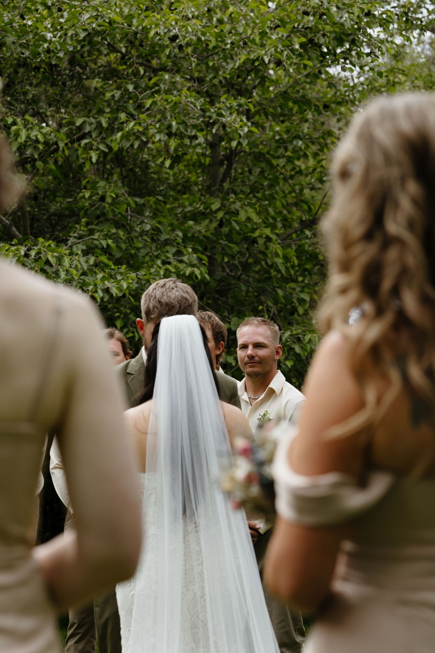 Guest smiles watching bride and groom kiss at outdoor wedding ceremony.