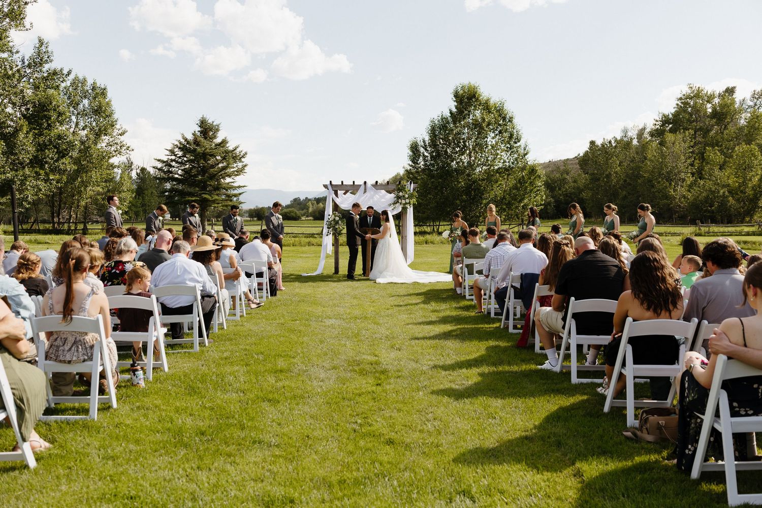 Outdoor summer wedding ceremony on green lawn with white chairs and mountain backdrop.