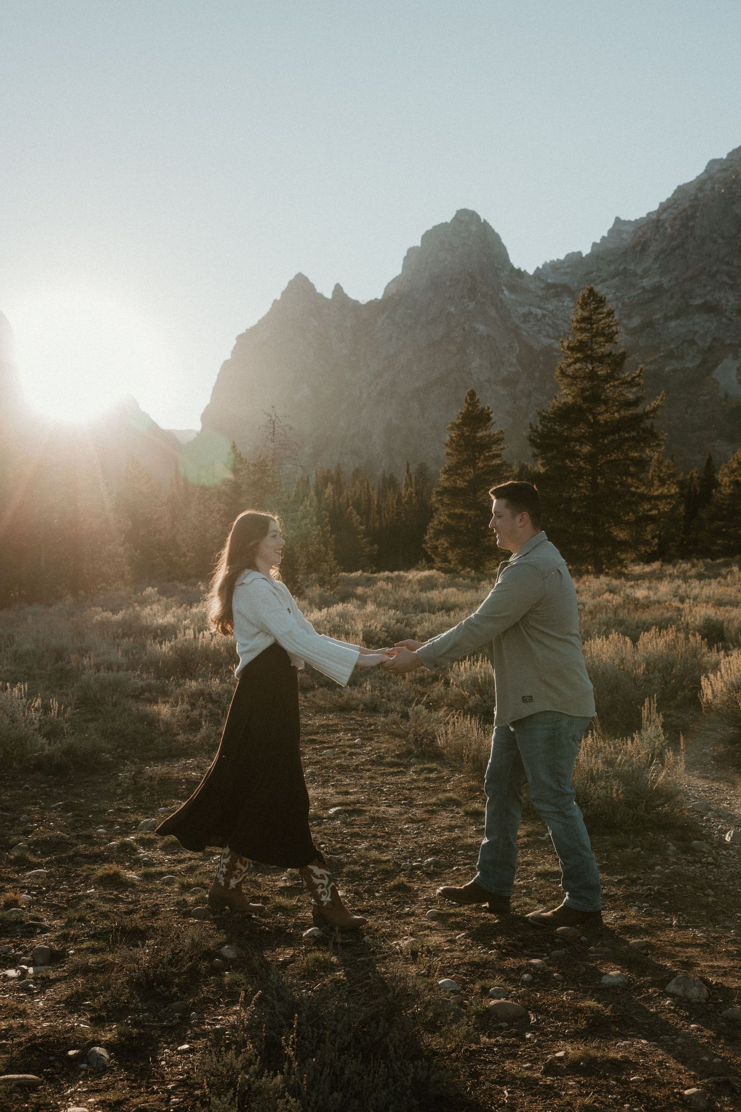 Couple holding hands and dancing in a sunlit mountain meadow with pine trees and rocky peaks at sunset.