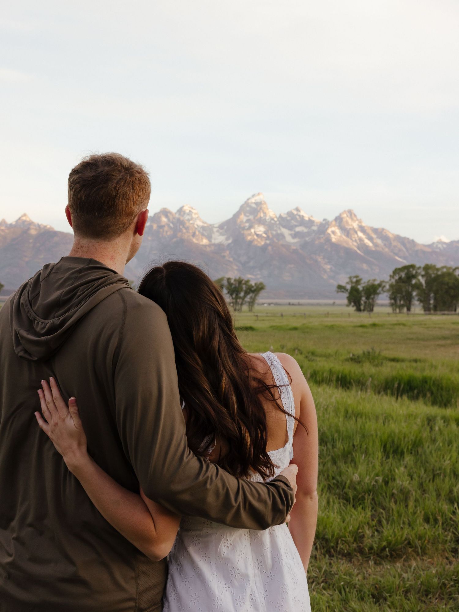 Couple with arms around each other gazing at the Grand Teton mountain range across a green meadow.