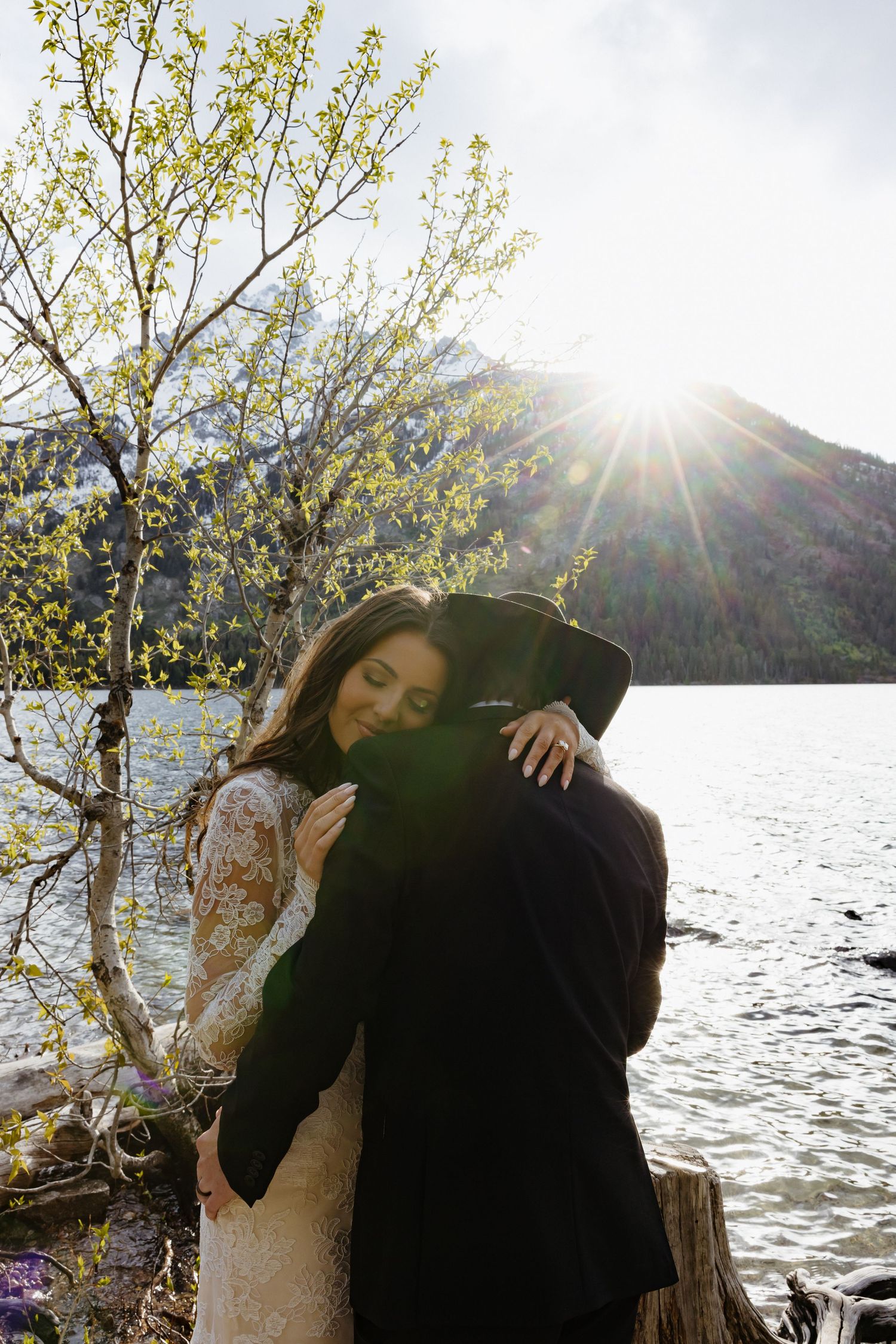 Bride and groom embracing by a sunlit mountain lake, surrounded by spring foliage and scenic wilderness.