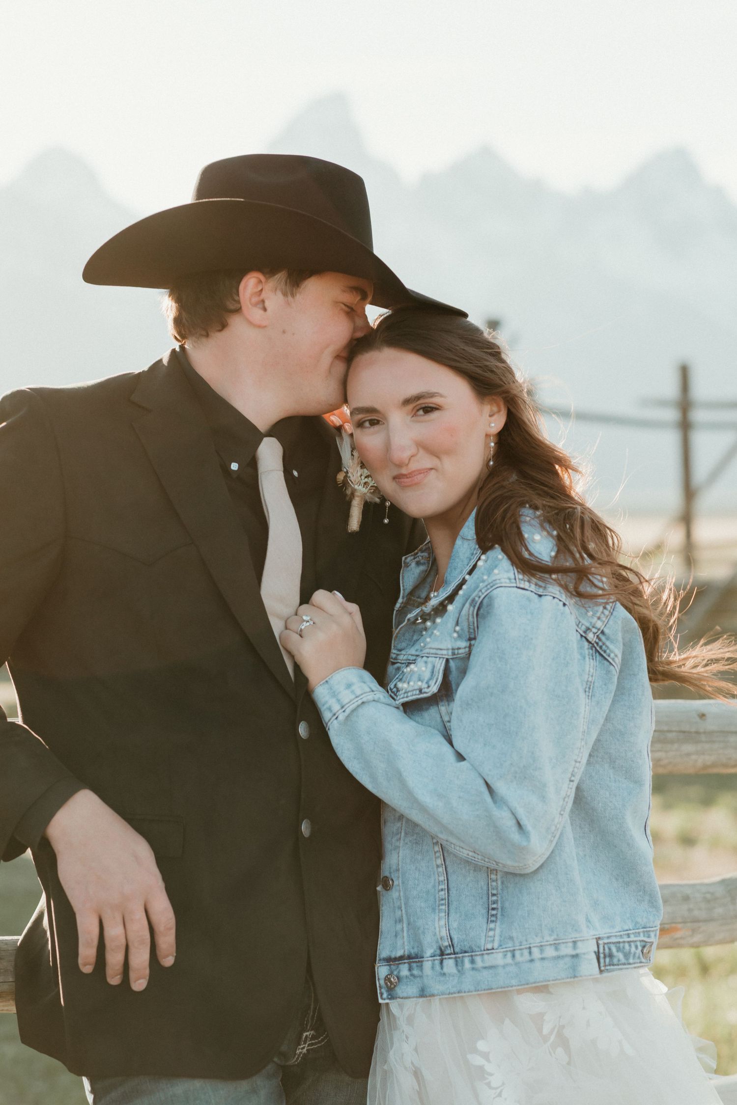 Groom in black cowboy hat kisses bride wearing denim jacket near fence at golden hour.