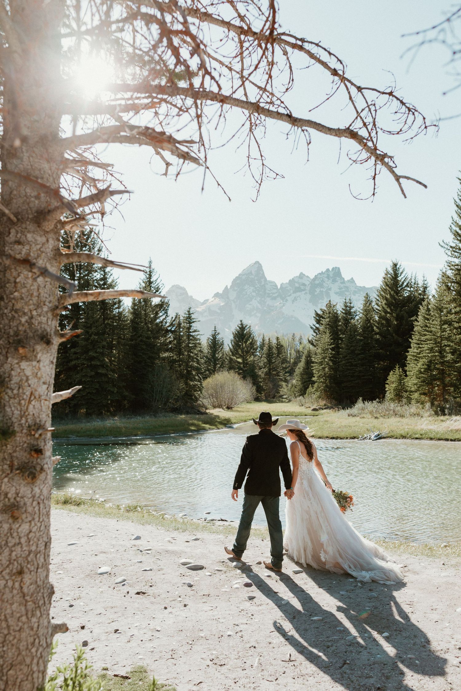 Bride and groom walk hand in hand toward a scenic mountain lake surrounded by pine trees on their wedding day.