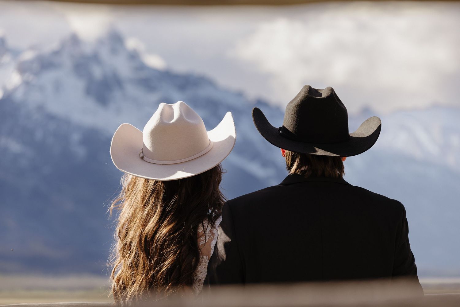 Bride and groom in matching white and black cowboy hats sit together admiring snow-capped Teton mountains.