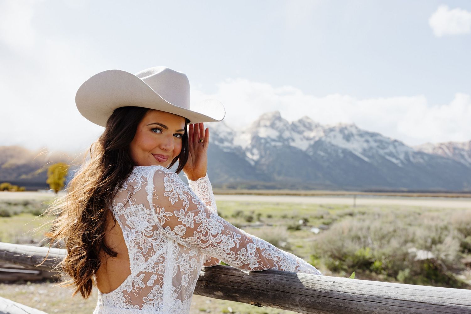 Beautiful bride in lace wedding gown and white cowboy hat poses by rustic fence with snowy Teton mountains behind.