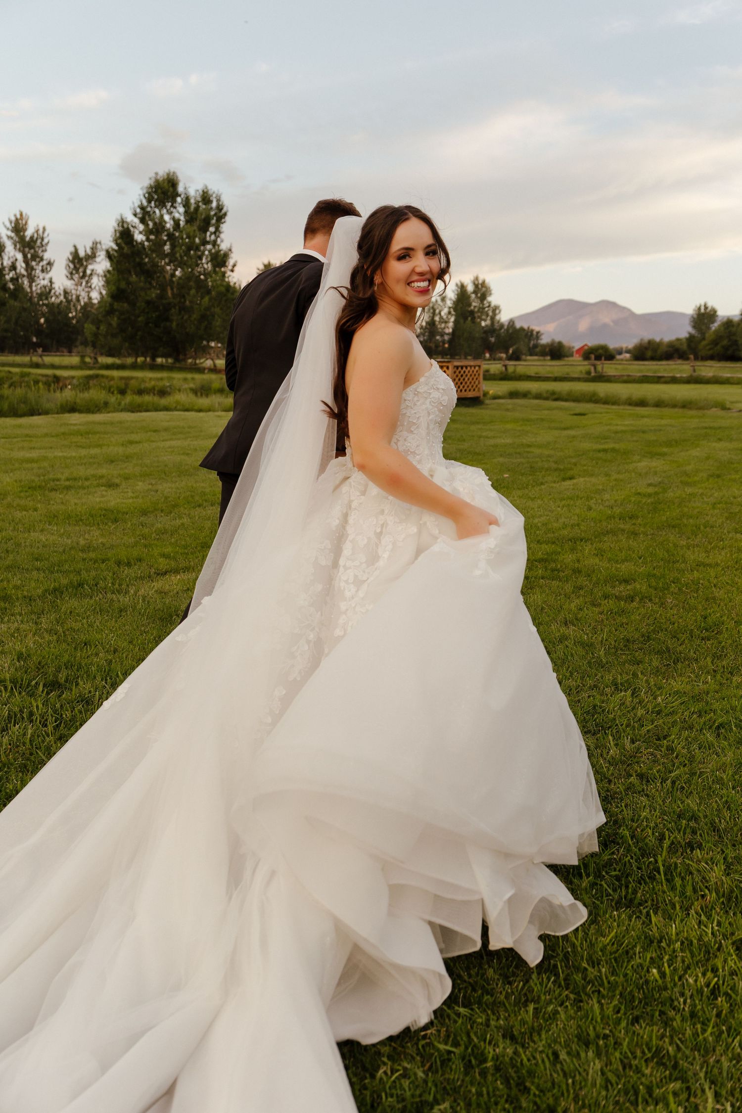 Smiling bride in lace ball gown walking with groom across green field at sunset with mountains behind.