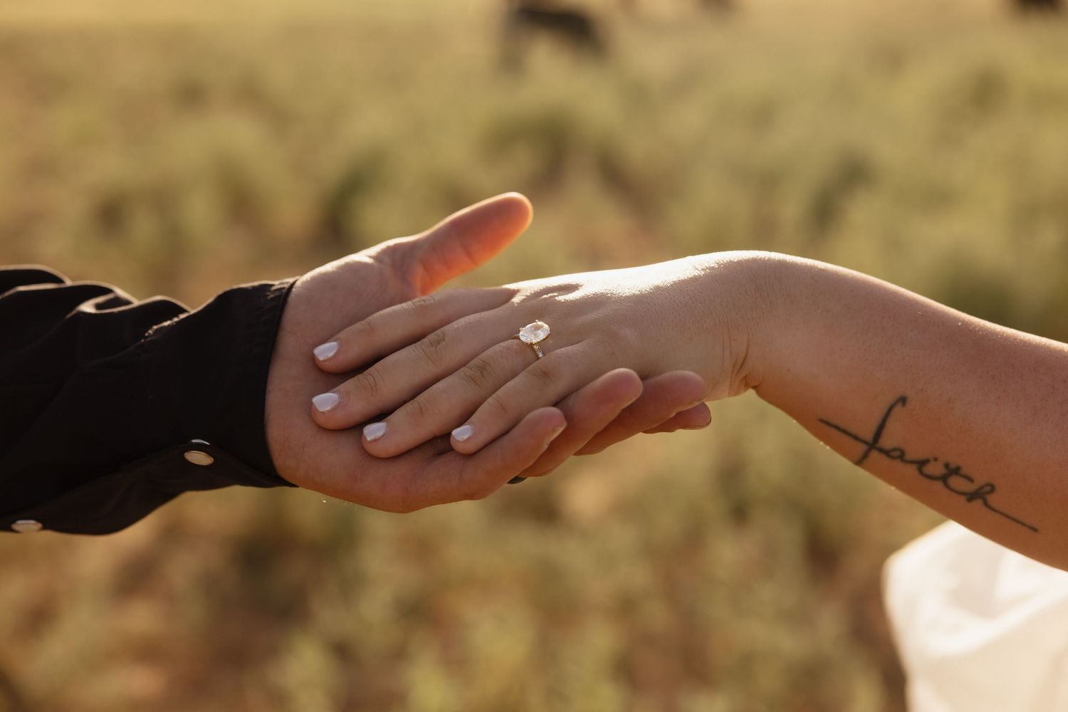 Close-up of two hands touching, one wearing an engagement ring, with a tattoo reading 'faith' on the wrist.