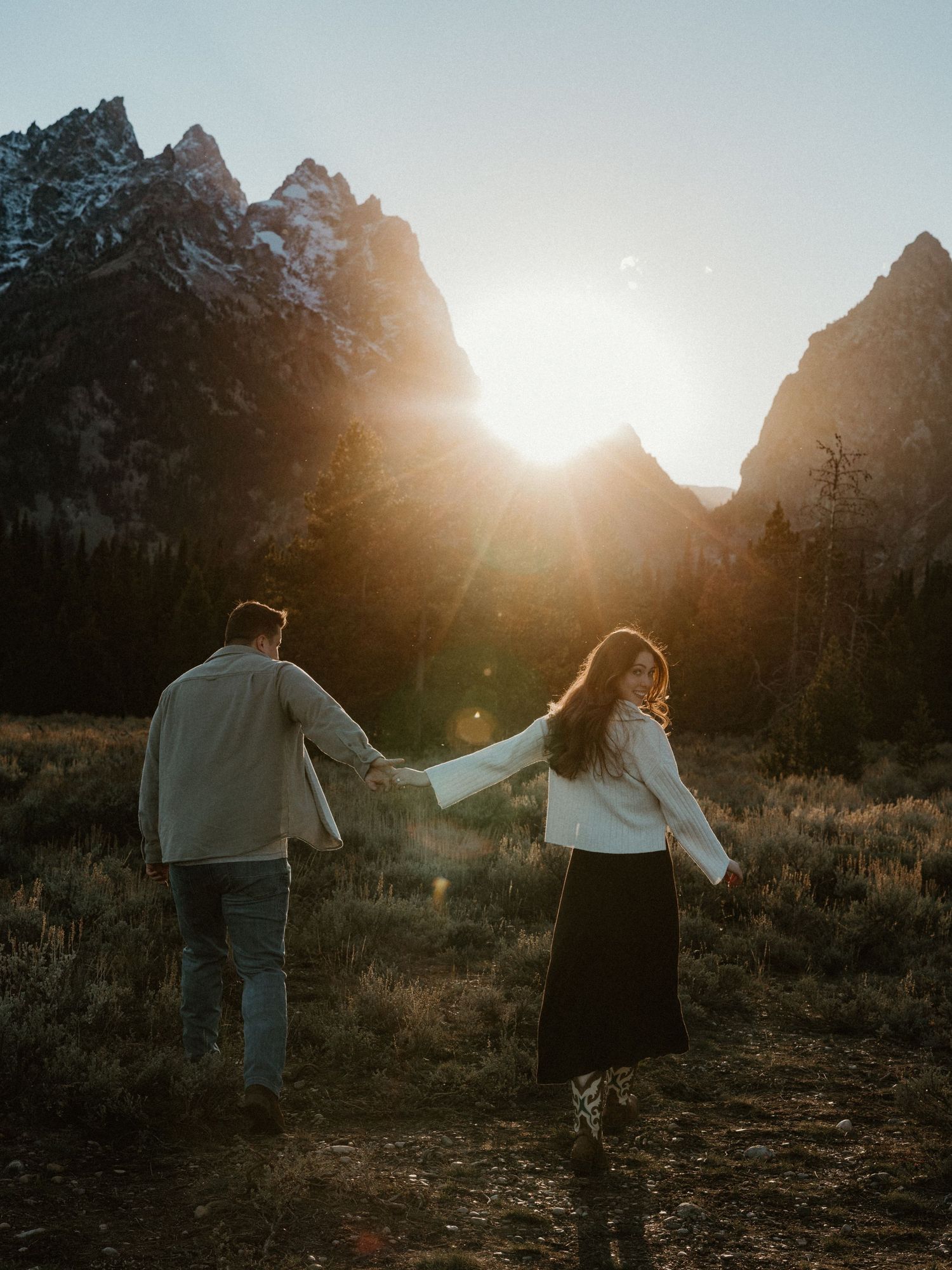 Couple holding hands walking toward brilliant sunset between towering rocky mountain peaks in a meadow.