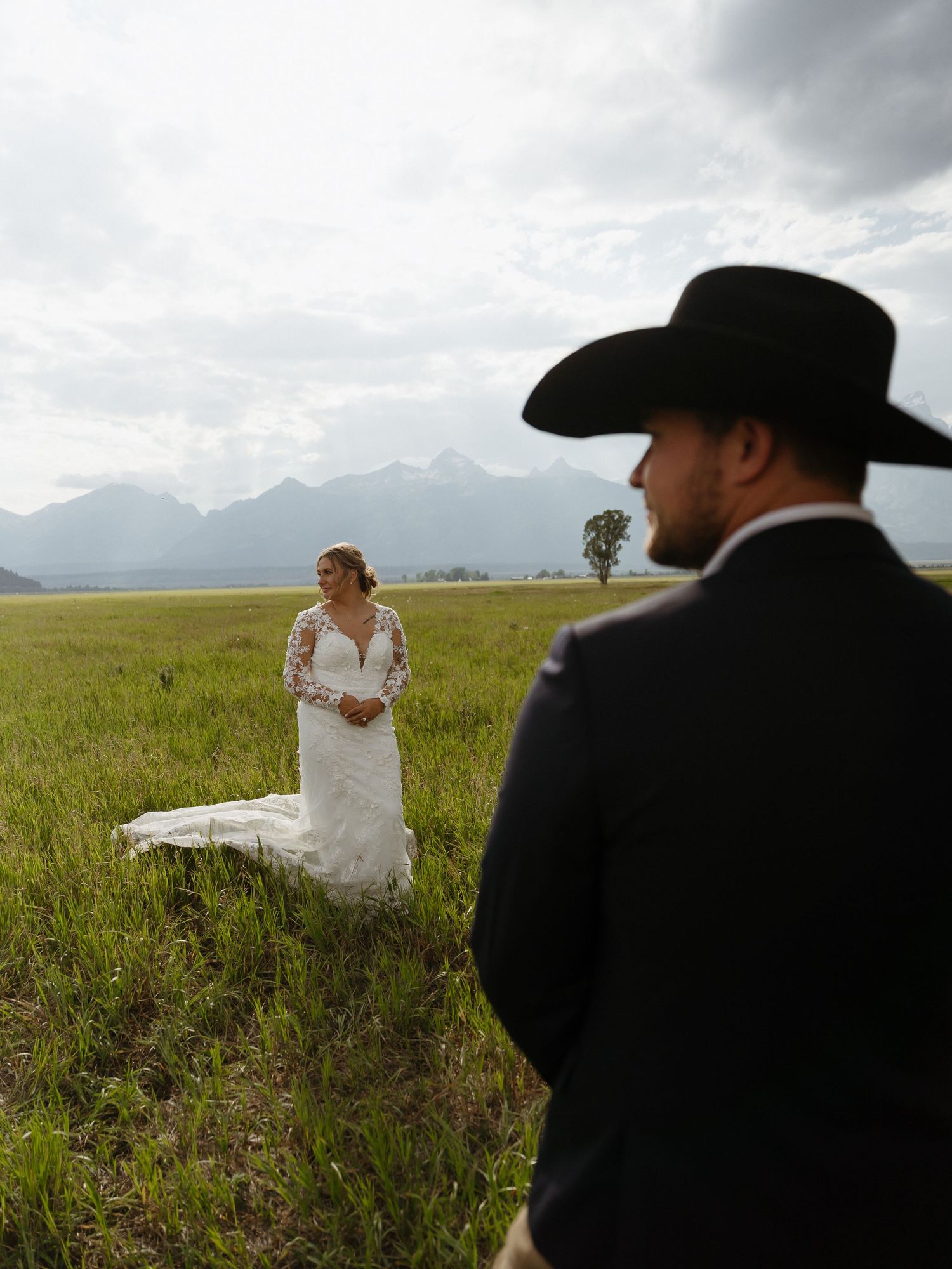 Bride in lace gown stands in green meadow as groom in black cowboy hat watches with Tetons behind.
