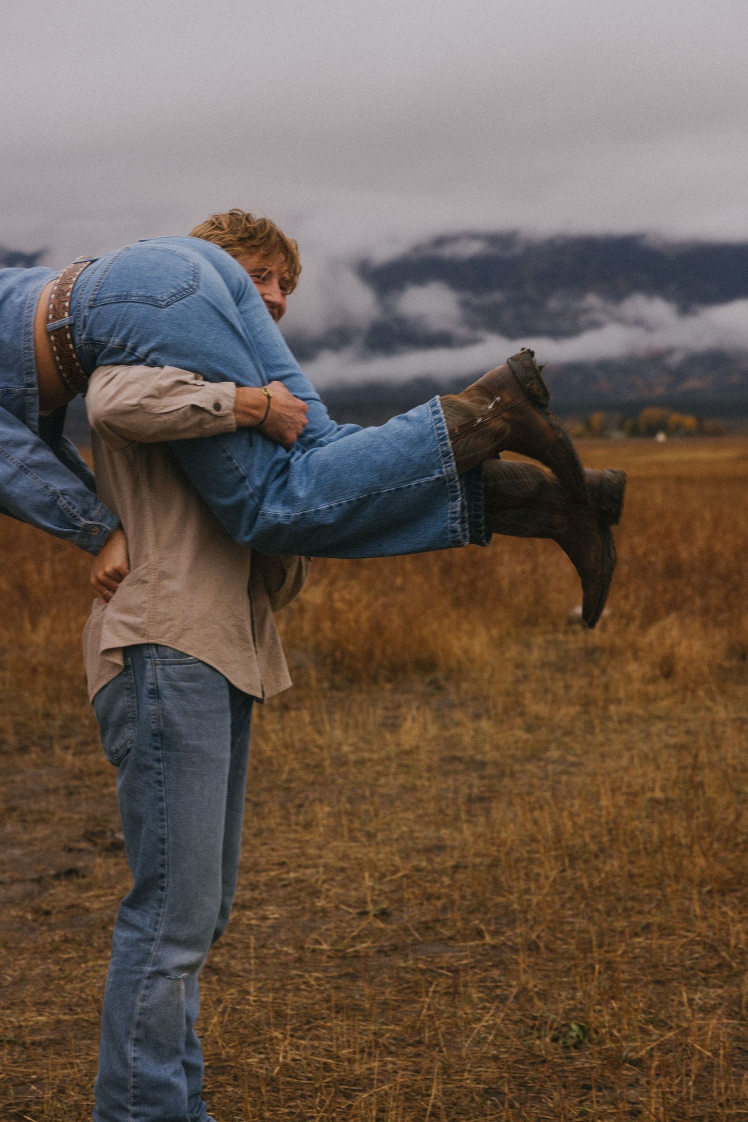 Man carrying woman over his shoulder in a golden autumn field with dramatic cloudy mountain backdrop.