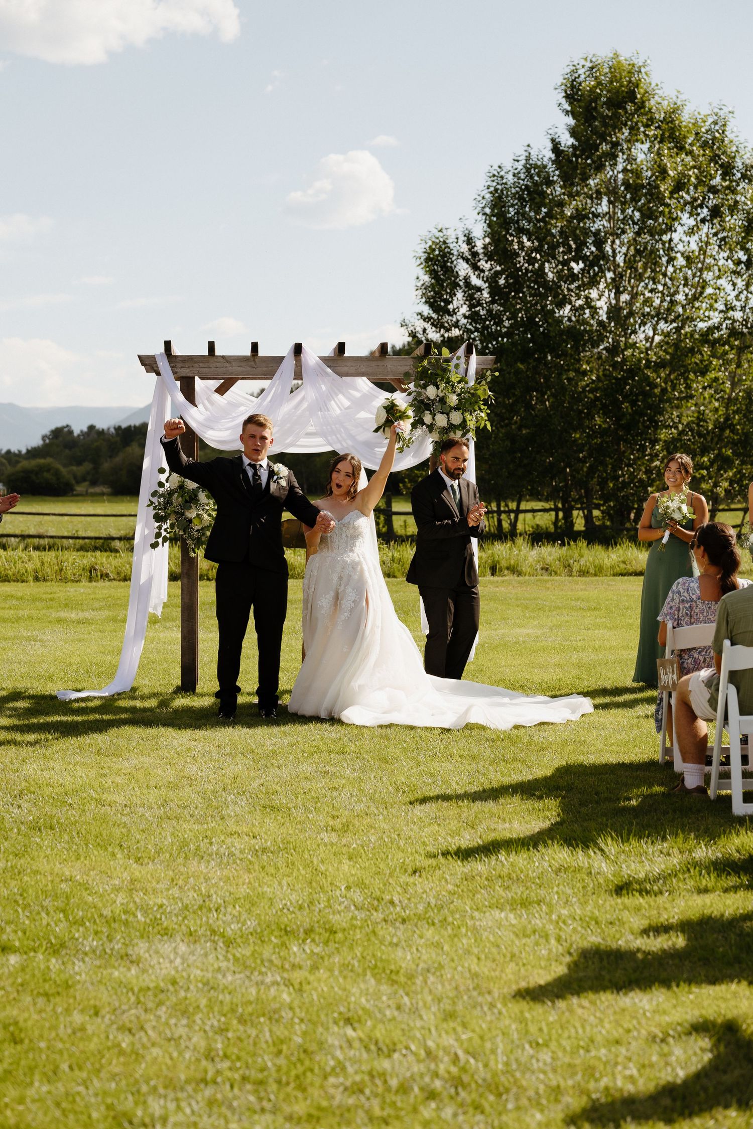 Newlyweds raise arms in celebration under floral wedding arch on sunny green lawn.