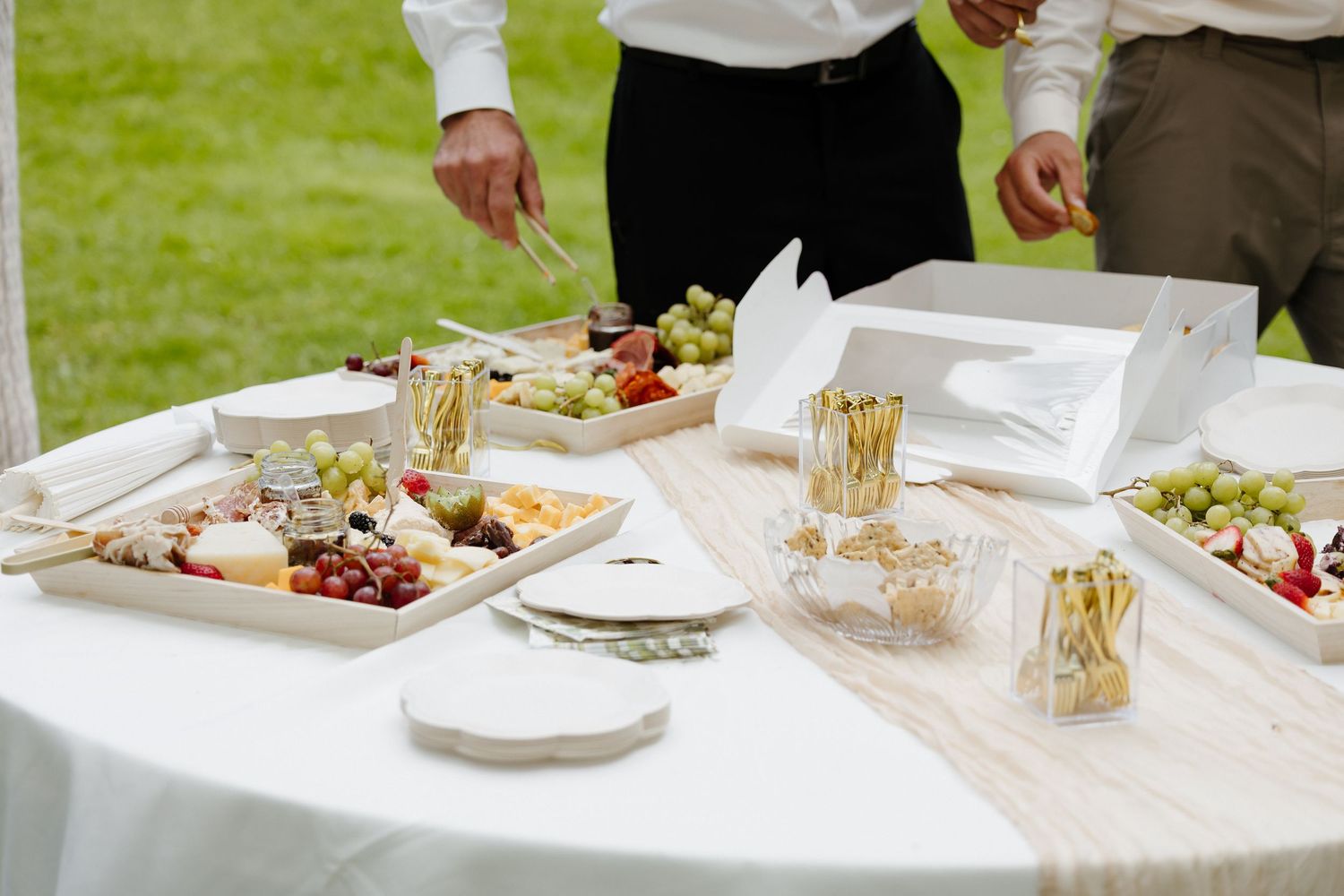 Elegant wedding reception appetizer table with charcuterie boards, fresh grapes, and golden glassware under a white tent.