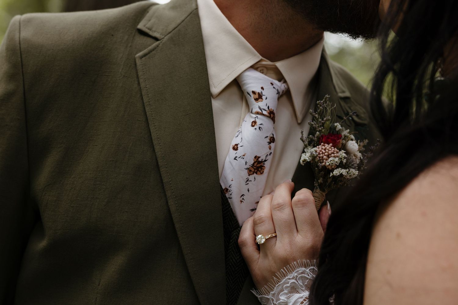 Close-up of groom in olive suit with floral tie and boutonniere, bride touching his lapel with ring.