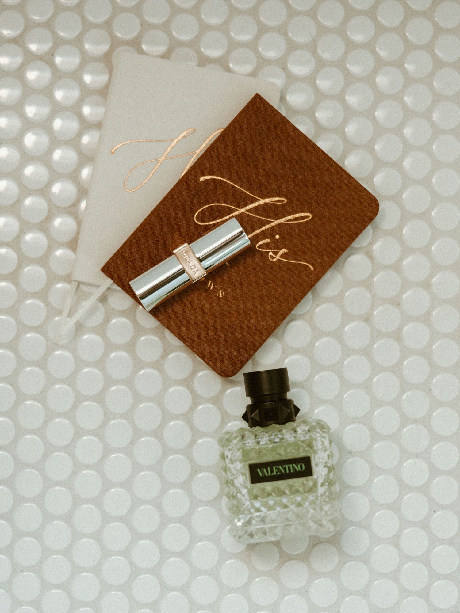 Valentino perfume bottle beside His and Hers vow books and lipstick on white tile background.