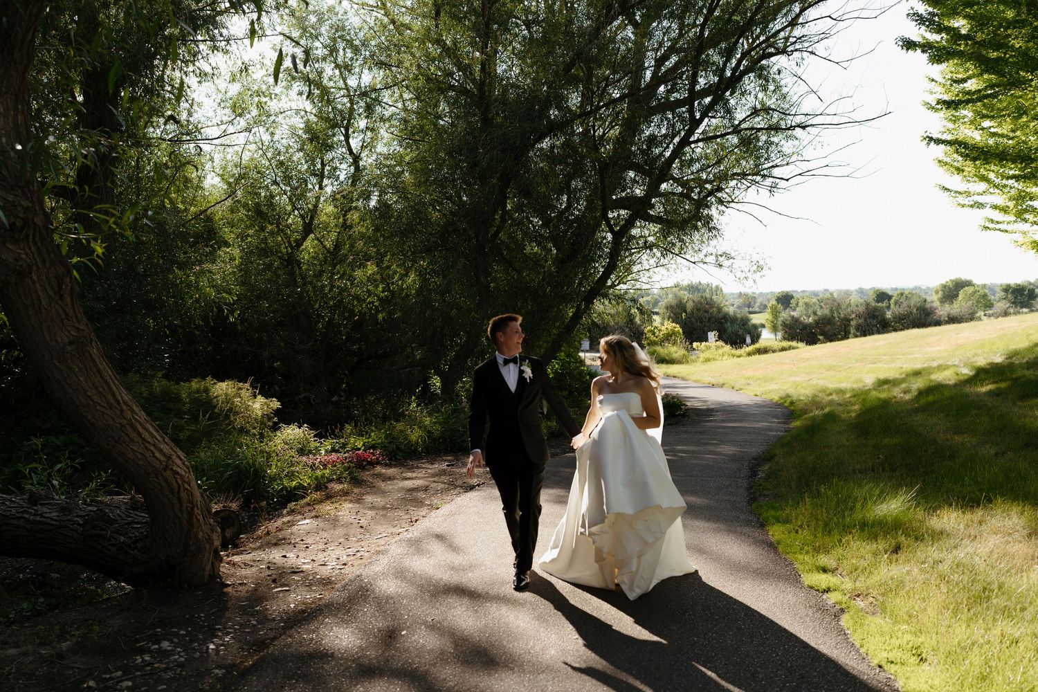 A bride and groom walk hand in hand along a sunlit winding path surrounded by lush green trees.