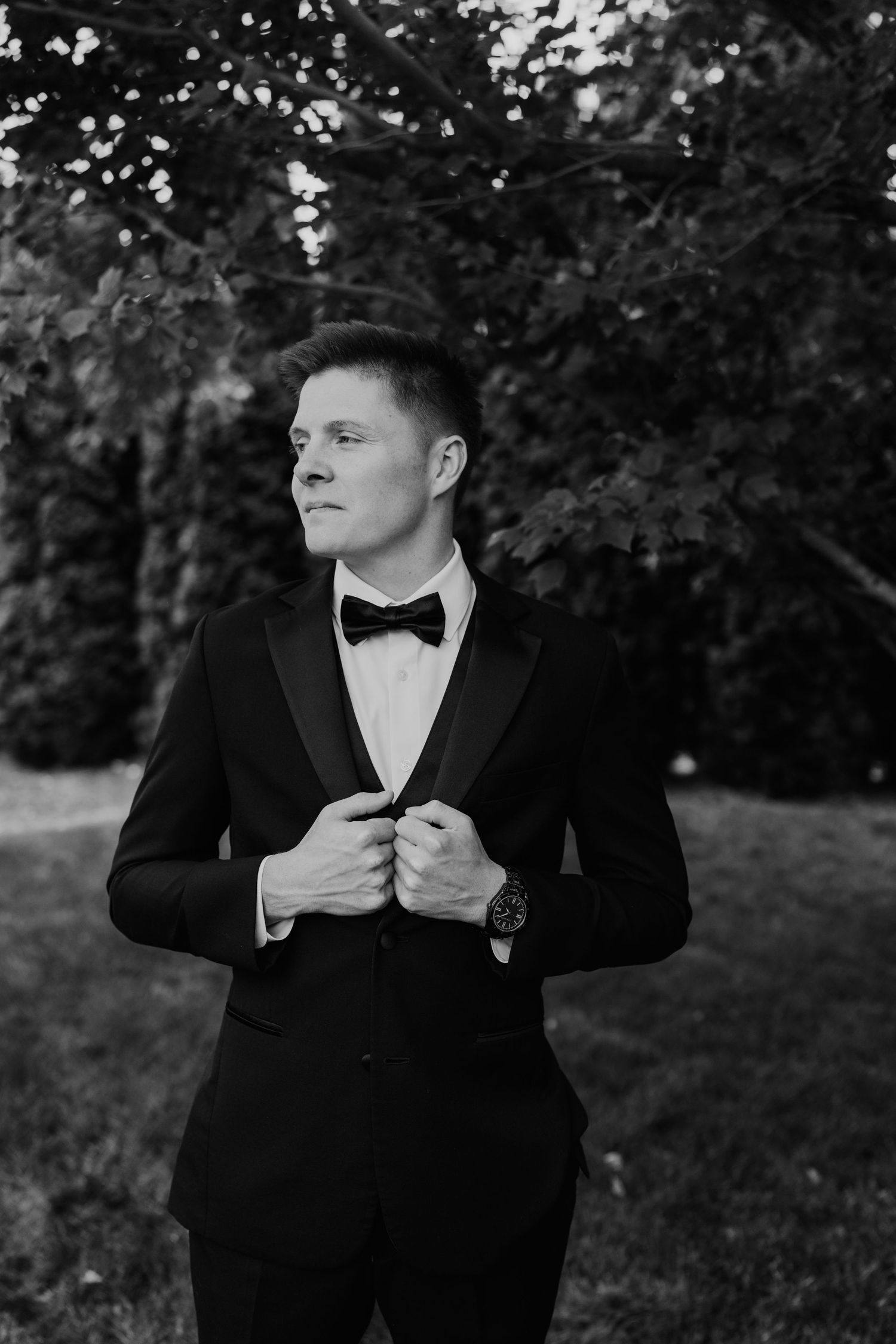 Elegant groom in black tuxedo with bow tie adjusting jacket outdoors in black and white photo.