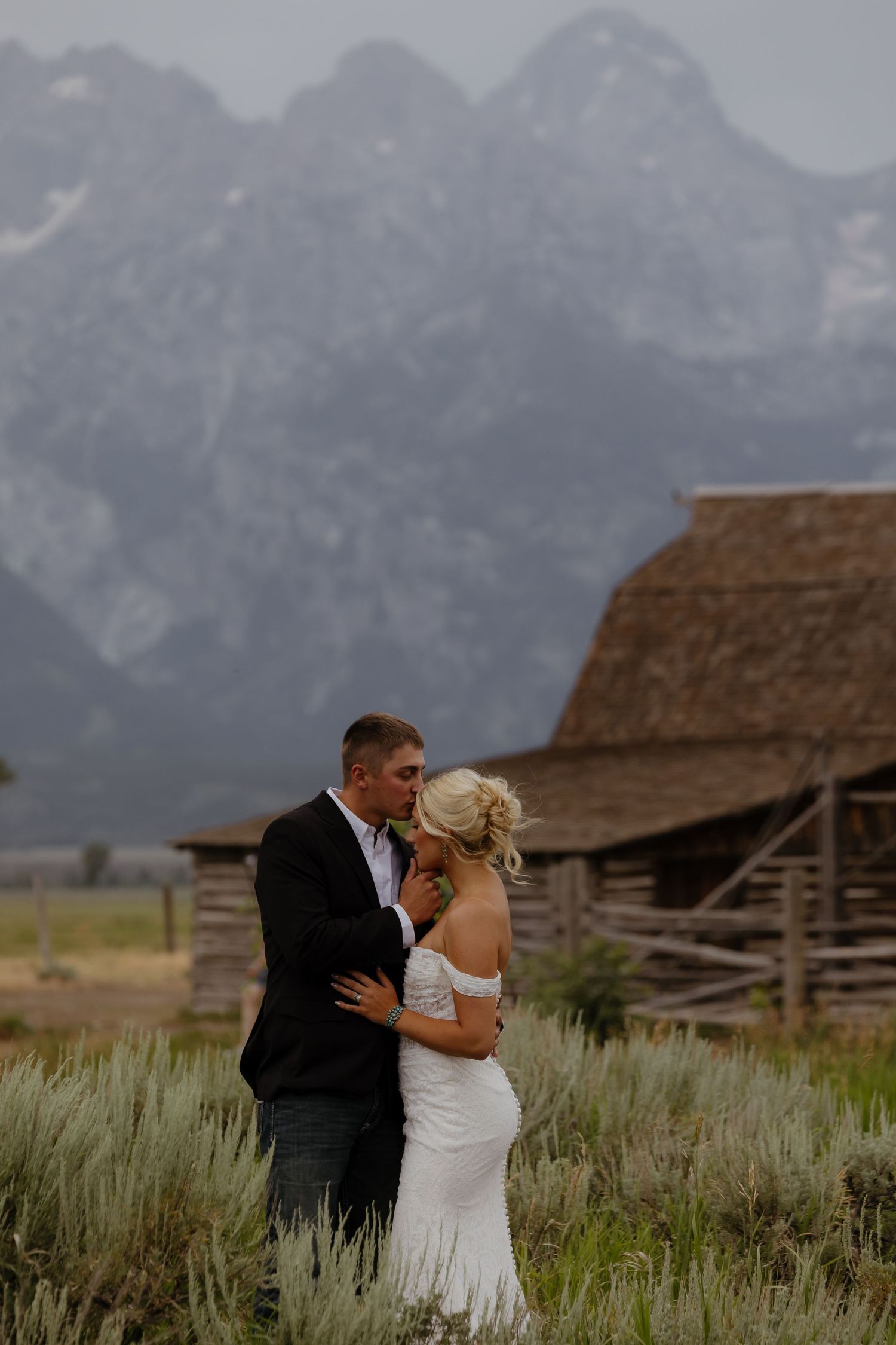 A bride and groom share a kiss in front of a rustic barn with the Grand Teton mountains in the background.