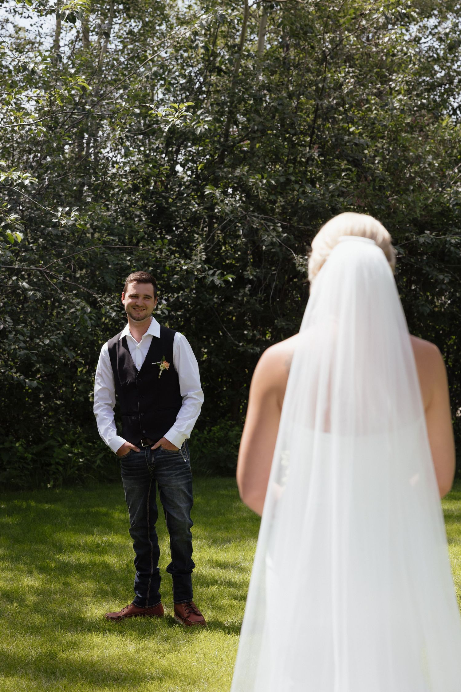 Groom smiling as bride in white veil approaches for their first look on wedding day outdoors.