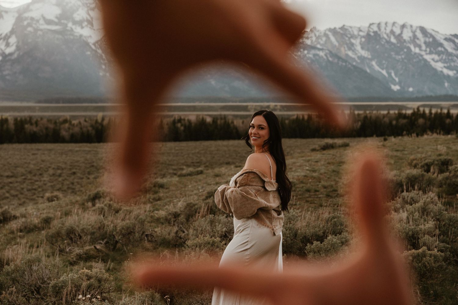 Woman smiling as hands frame her face like a camera with mountain meadow landscape in the background.
