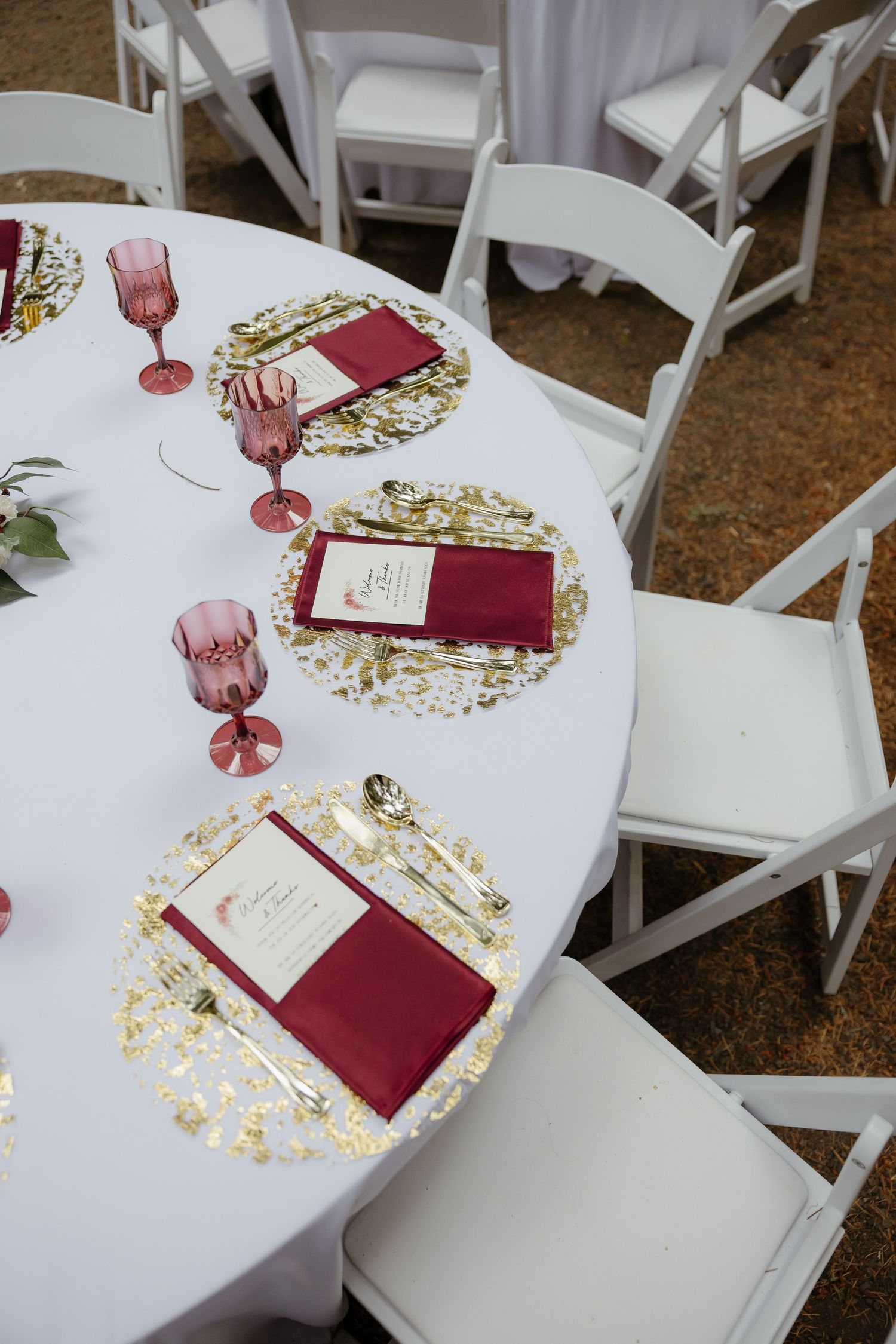 Elegant wedding table setting featuring gold chargers, burgundy napkins, pink glassware, and gold cutlery on white tablecloth.
