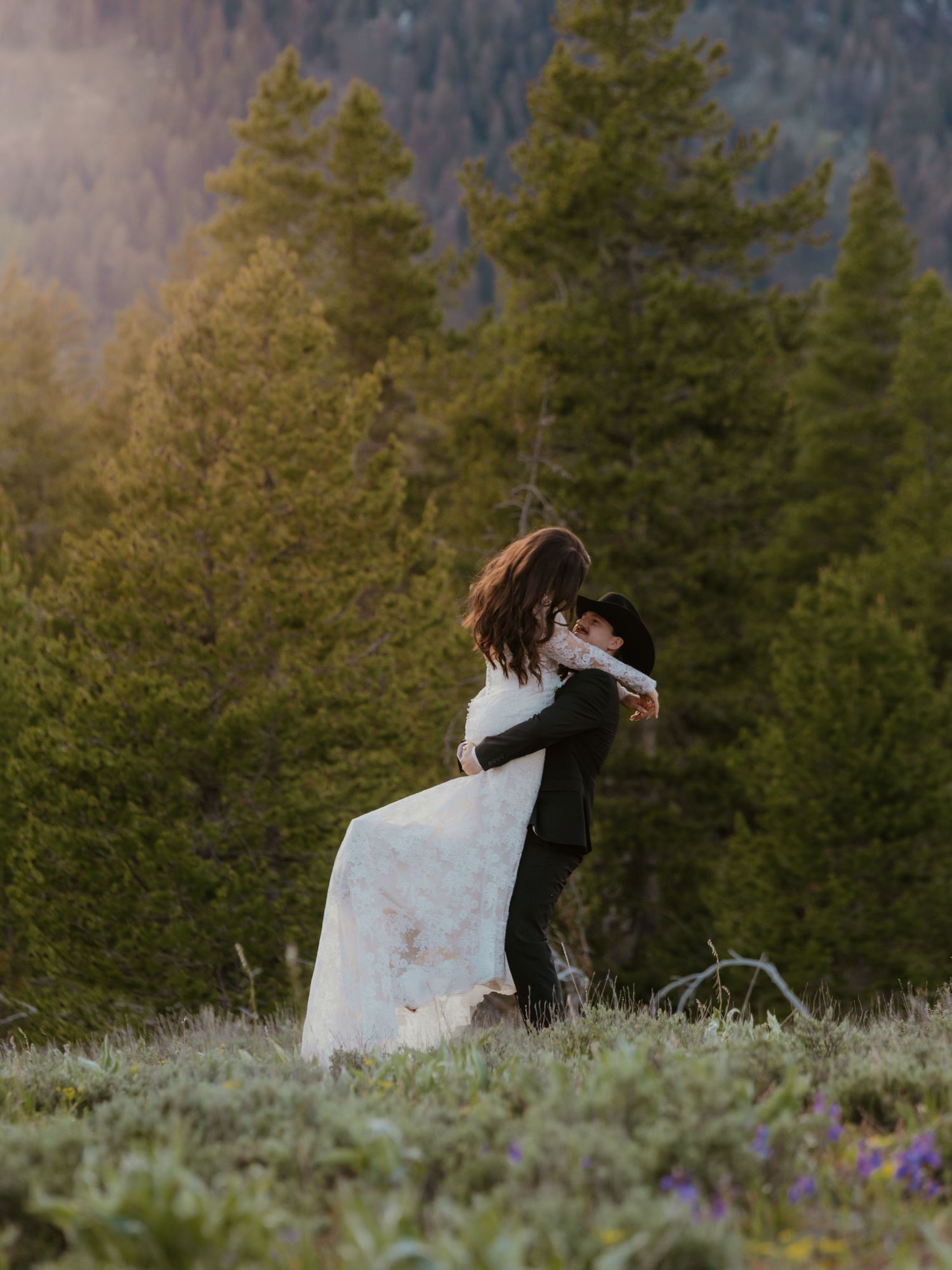 Groom lifting bride in a white gown in a wildflower meadow surrounded by pine trees at sunset.