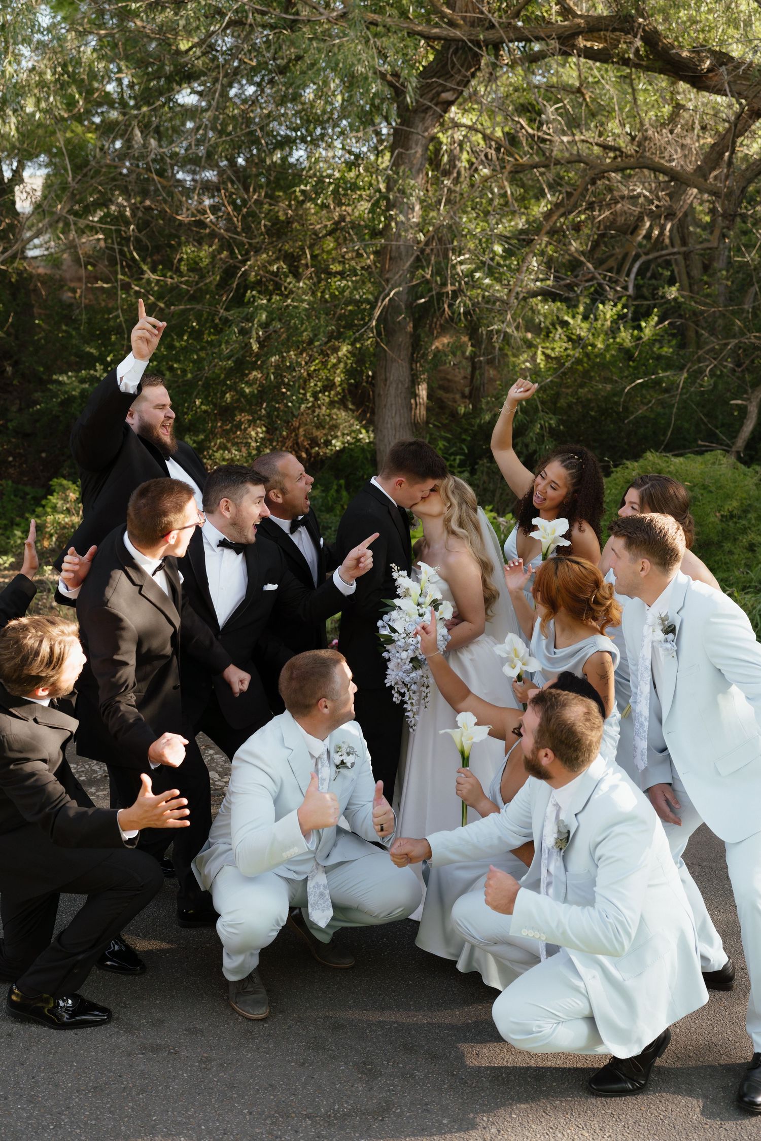 Wedding party celebrating outdoors with bride and groom surrounded by groomsmen in black tuxedos and white suits cheering joyfully.
