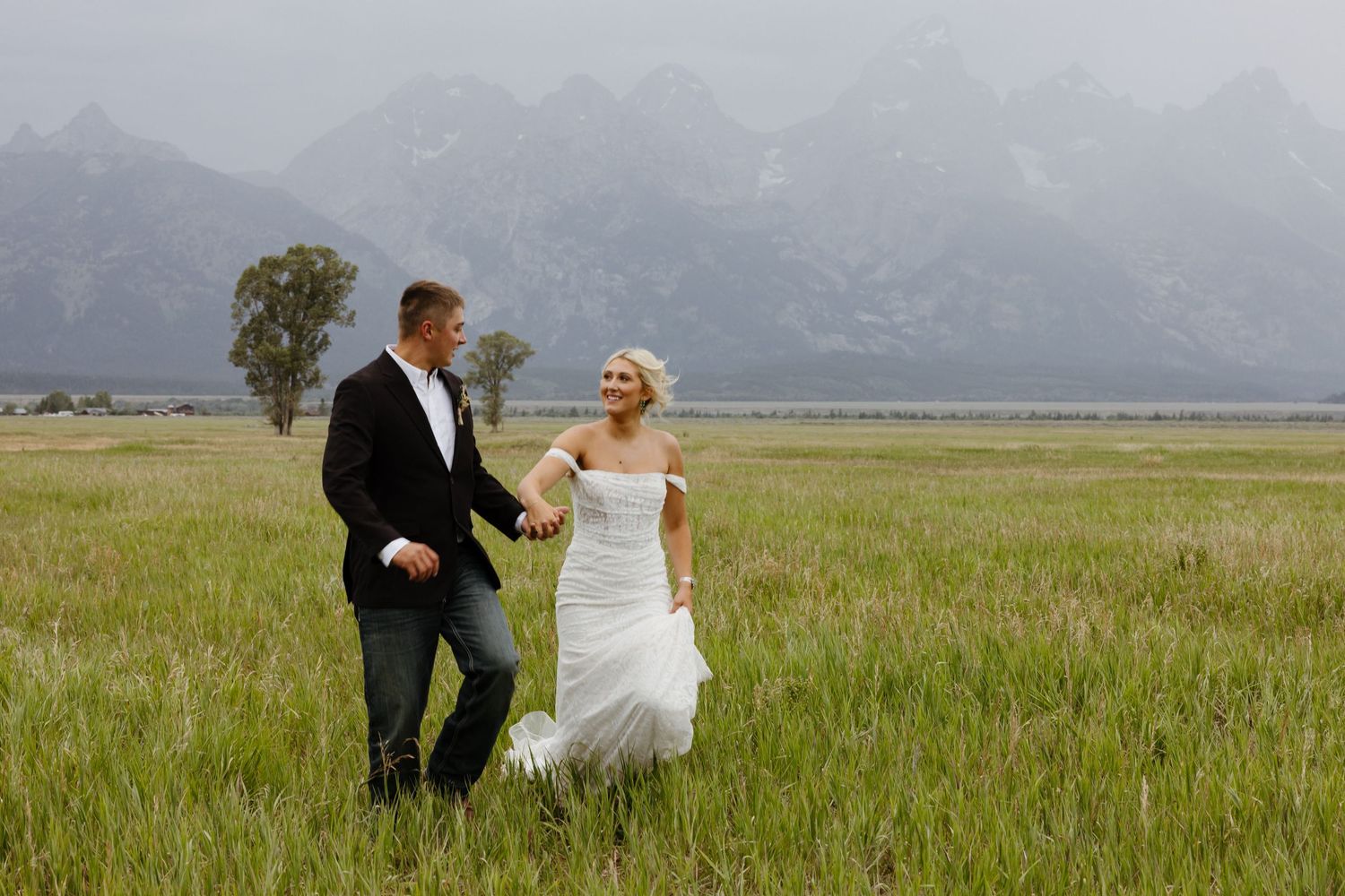 Bride and groom walking hand-in-hand through green meadow with Grand Teton mountains in background.