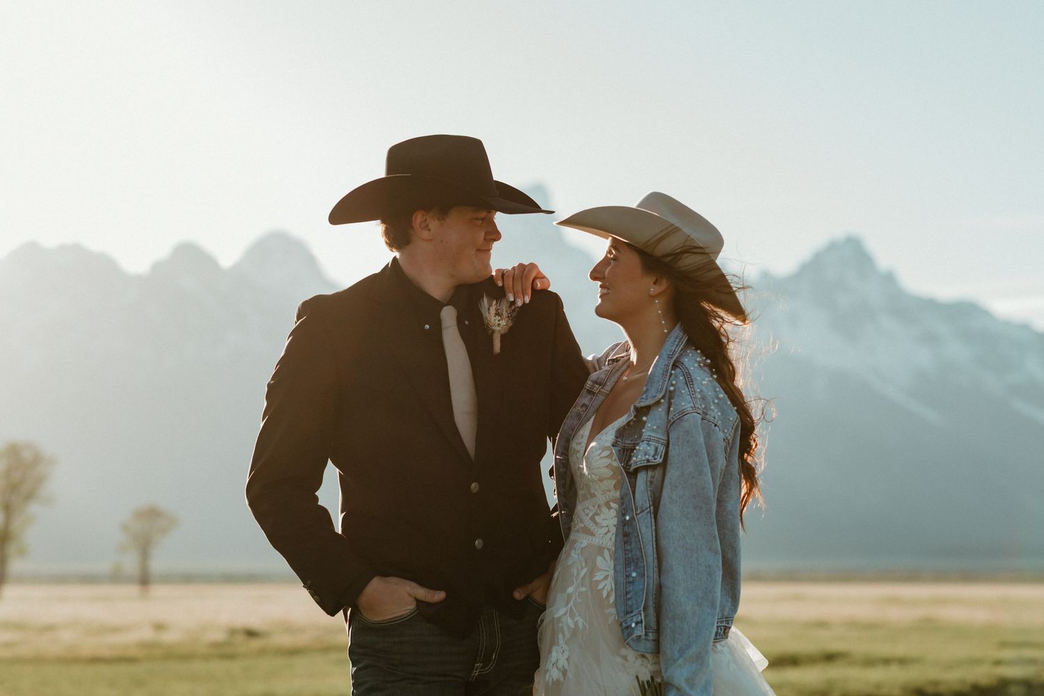 Western wedding couple in cowboy hats smile at each other with misty Teton mountain backdrop.