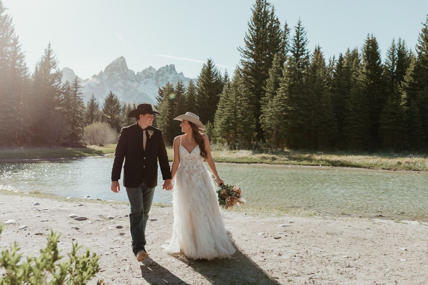 Bride in flowing lace gown and groom in cowboy hat walk together along a sunlit mountain river.