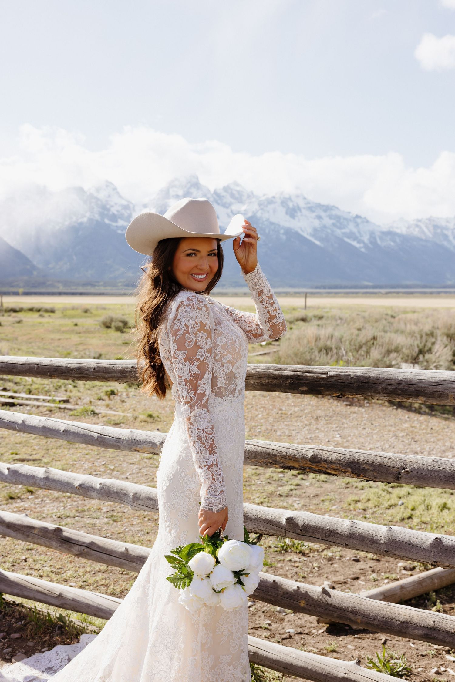 Smiling bride in lace mermaid gown tips white cowboy hat holding white peony bouquet with Teton mountains behind.