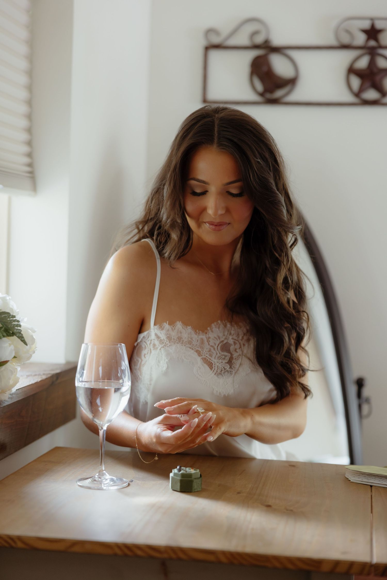 Bride in white lace slip dress examining her engagement ring at a wooden table with a glass of champagne.