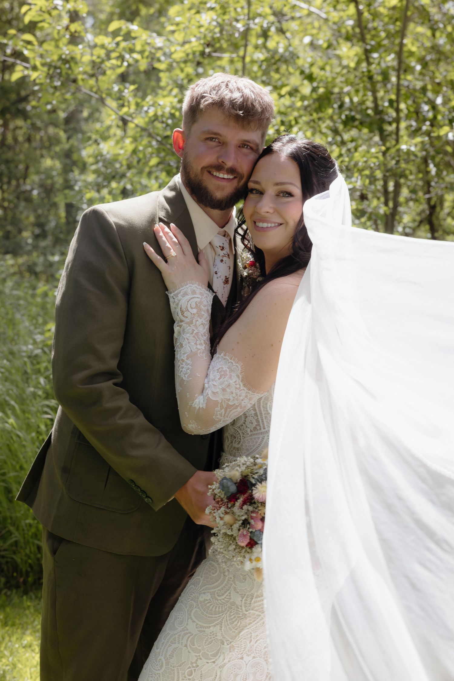 Smiling bride and groom portrait outdoors with flowing veil, lace dress, and olive suit in green setting.
