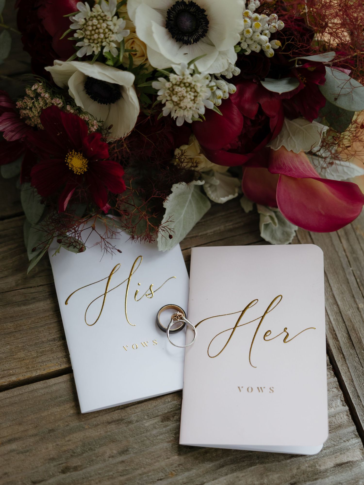 His and Her vows booklets with wedding rings beside lush burgundy and white floral bouquet.