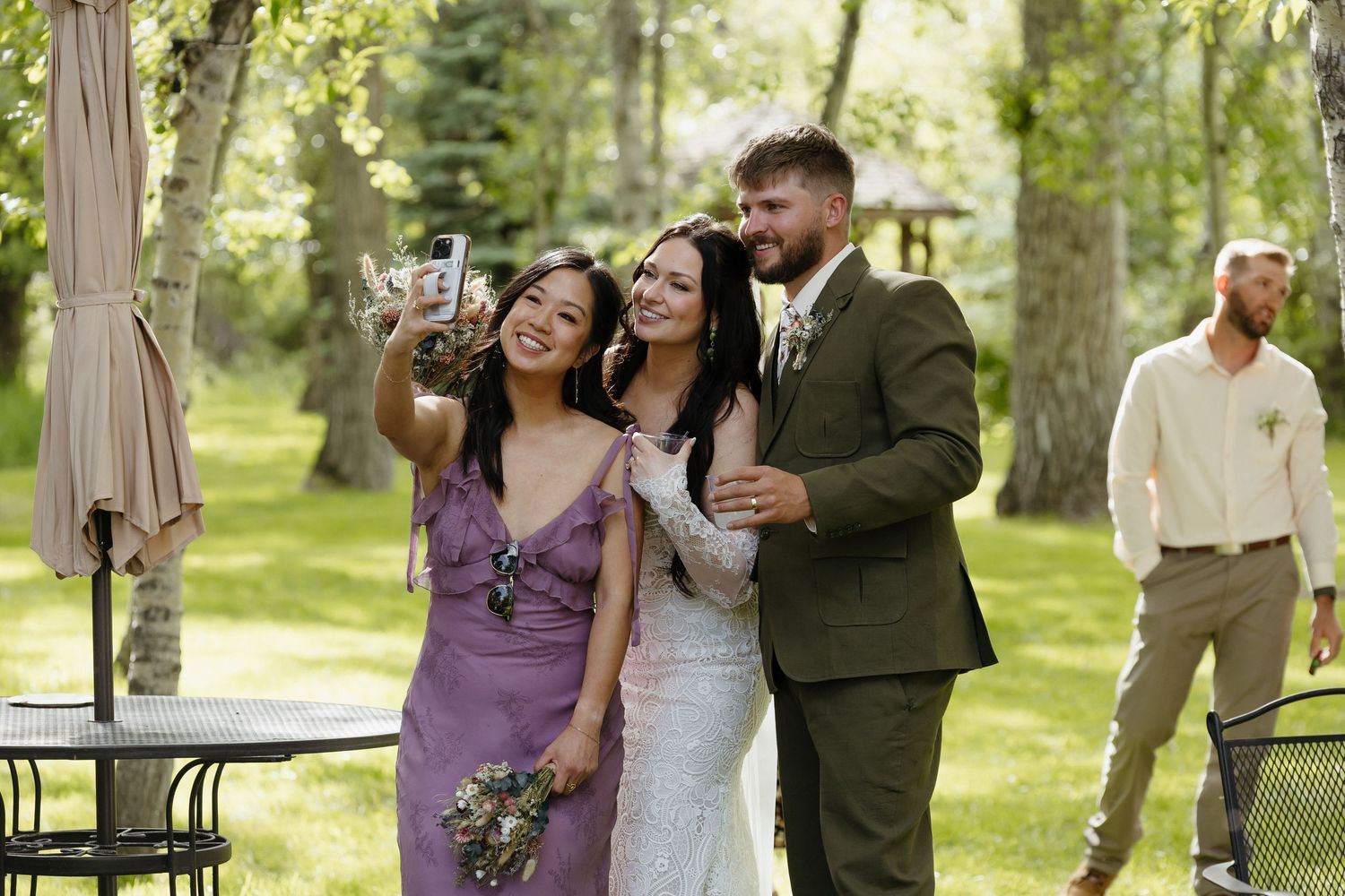 Bride and groom posing for selfie with bridesmaid in purple dress at outdoor wedding reception in wooded setting.