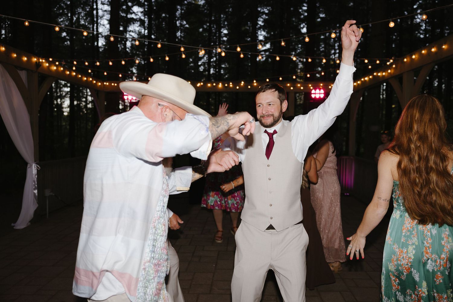 Groom in tan suit dances energetically with guest in white cowboy hat under string lights at forest reception.