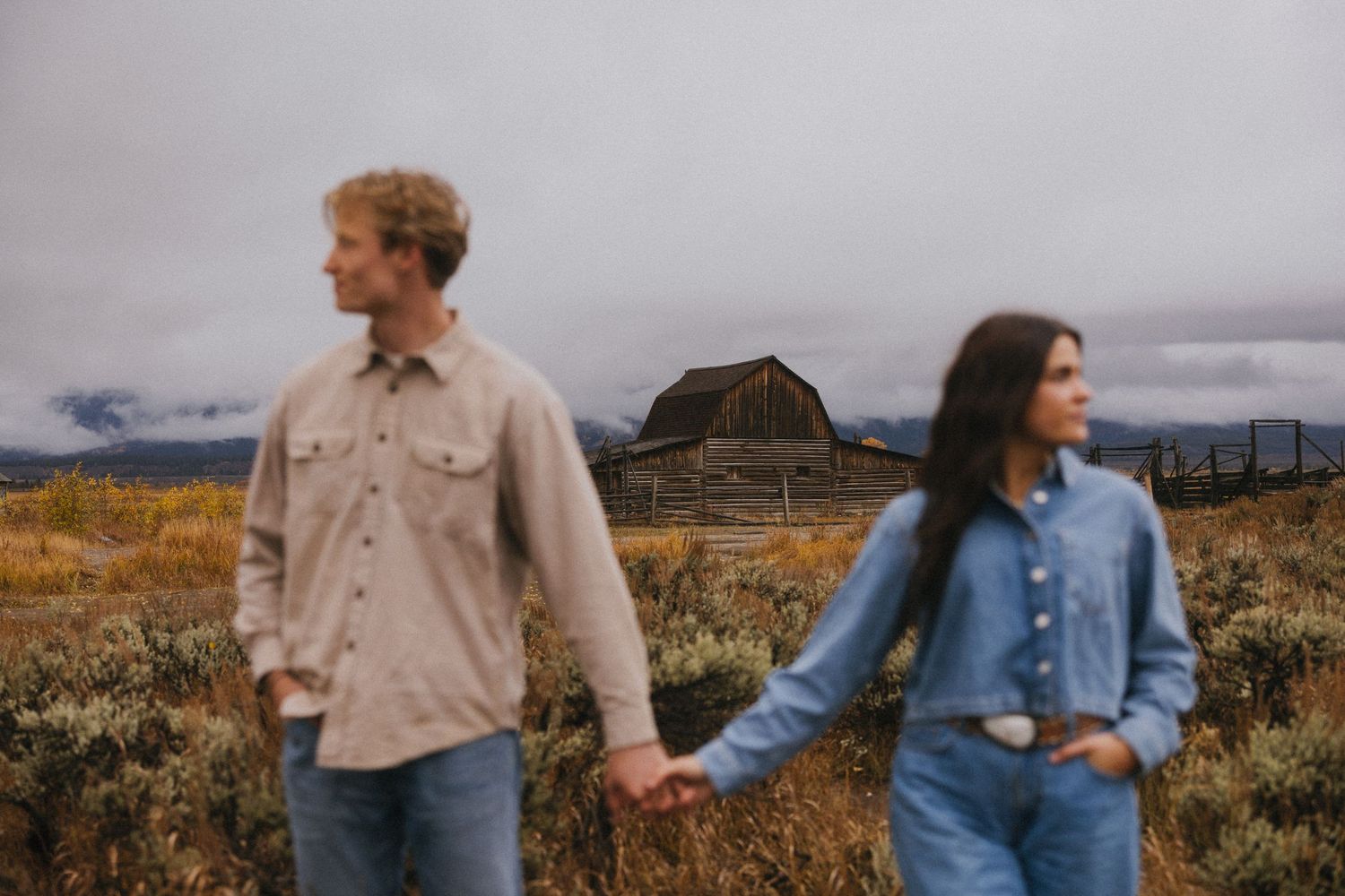 Couple holding hands walking through a golden field with a historic barn and cloudy mountains behind.