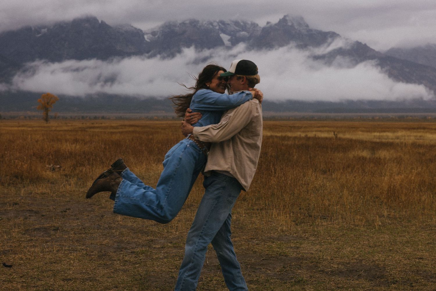 Man lifting woman in his arms while embracing in a golden autumn field with misty mountains.