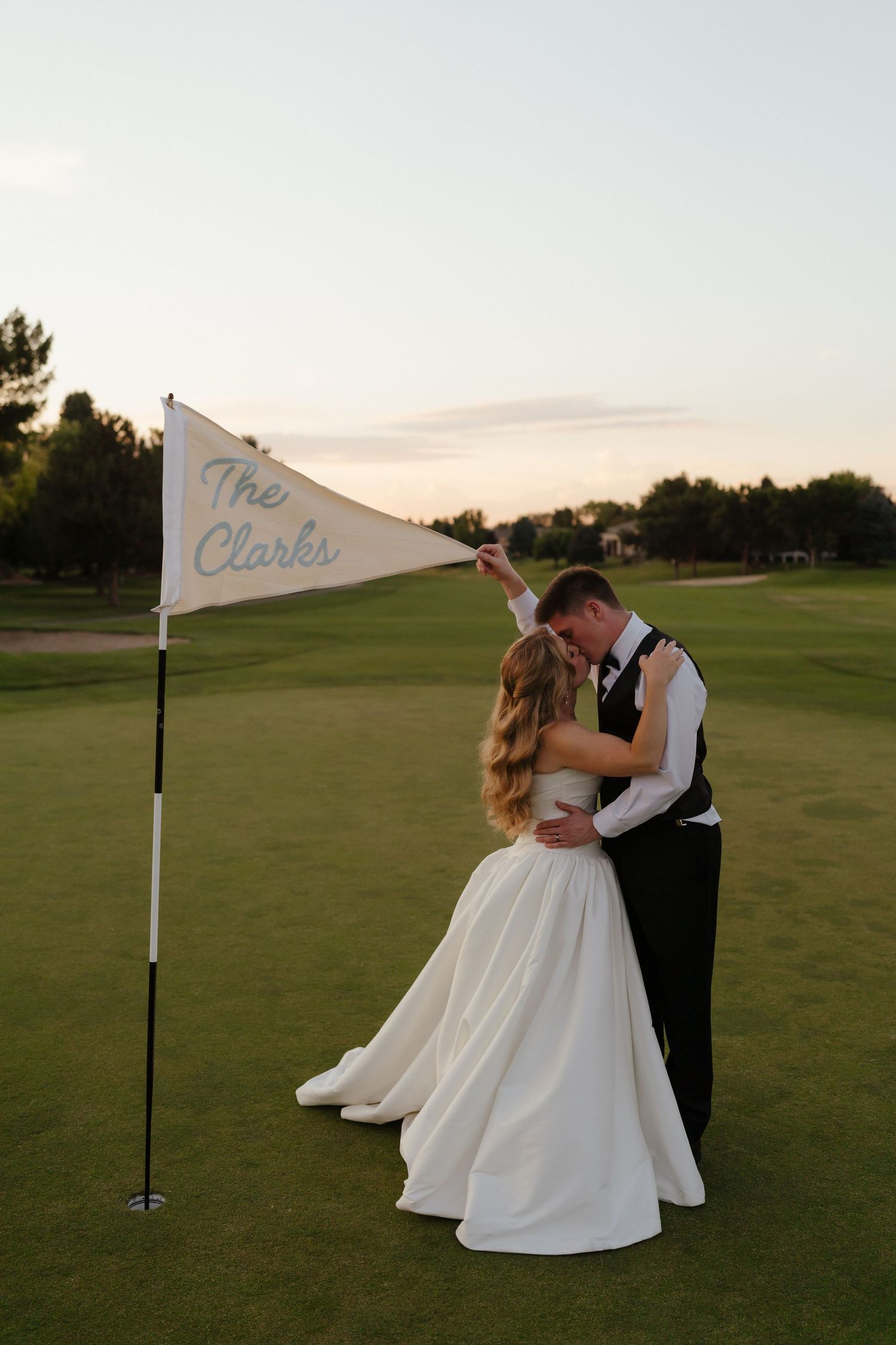Newlyweds kiss on golf course green beside a personalized pennant flag reading The Clarks at sunset.