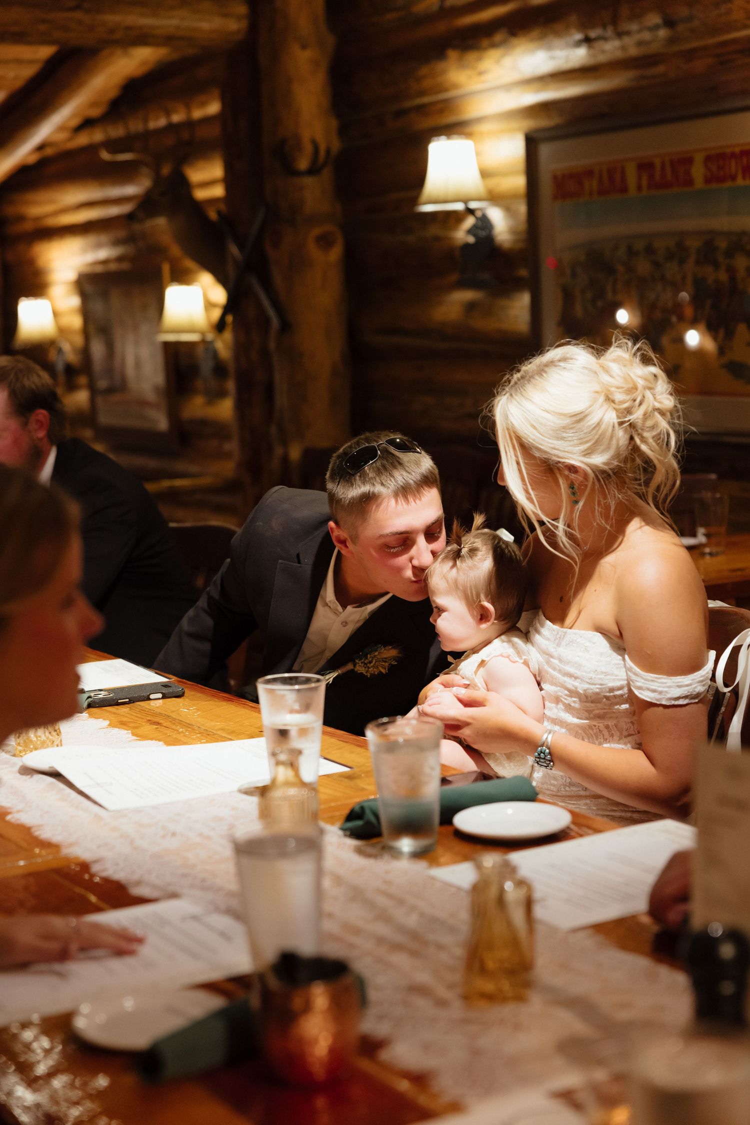 Bride and groom sharing a tender moment with a baby at a rustic log cabin wedding reception dinner table.