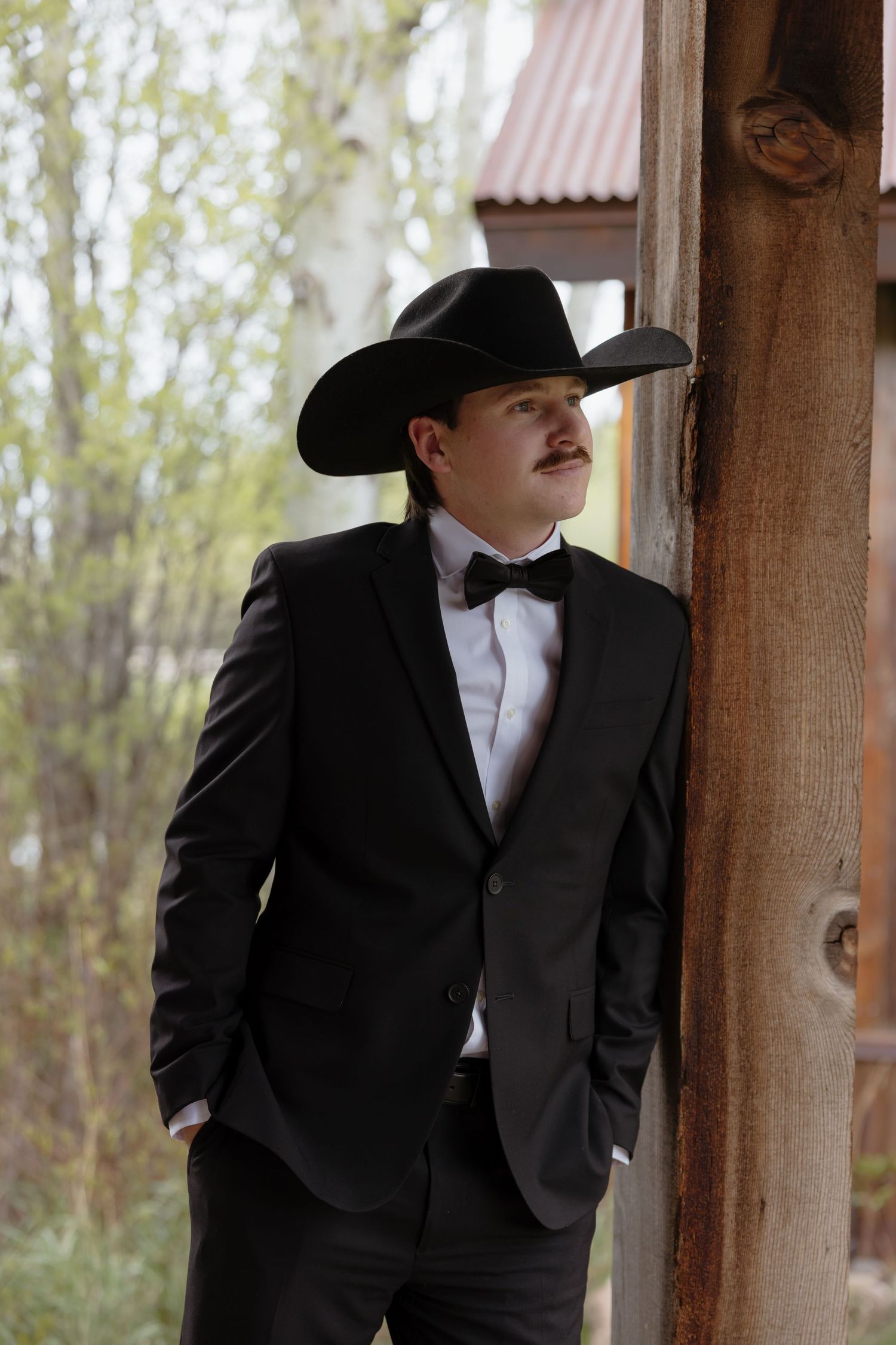 Groom in black tuxedo and cowboy hat leaning against a wooden post outdoors at a rustic venue.