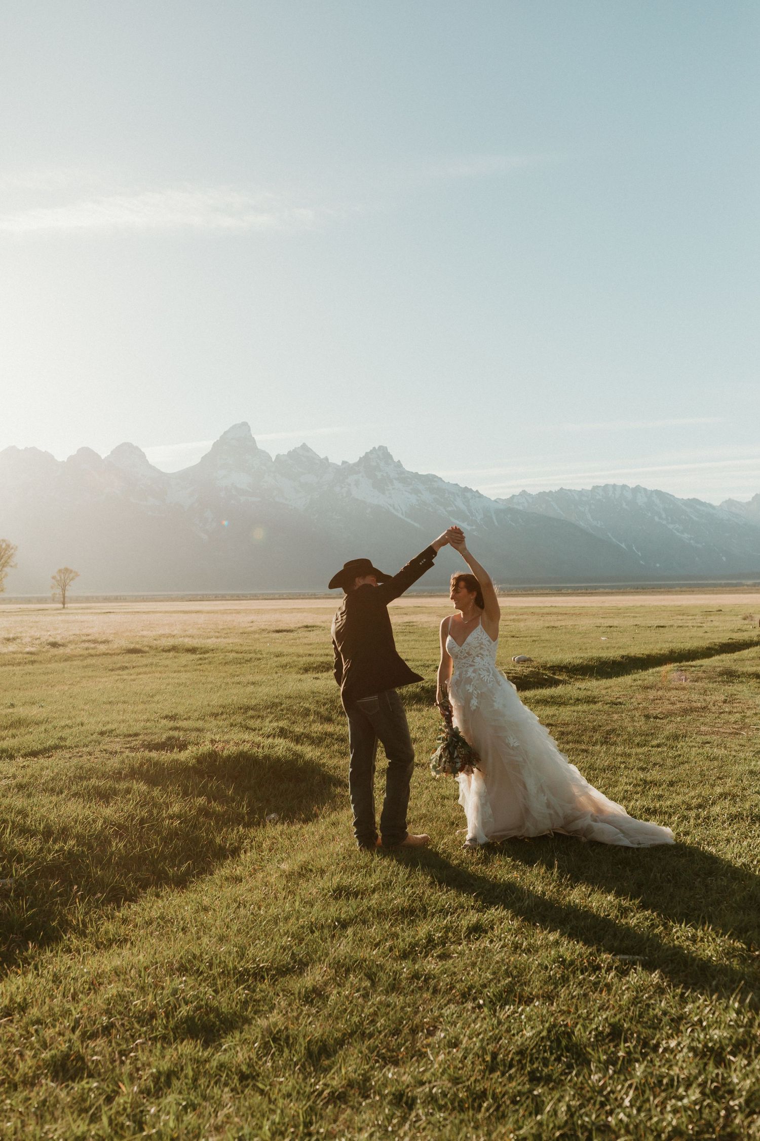 Groom twirls bride in flowing white gown on vast green meadow with Grand Teton mountains at sunset.