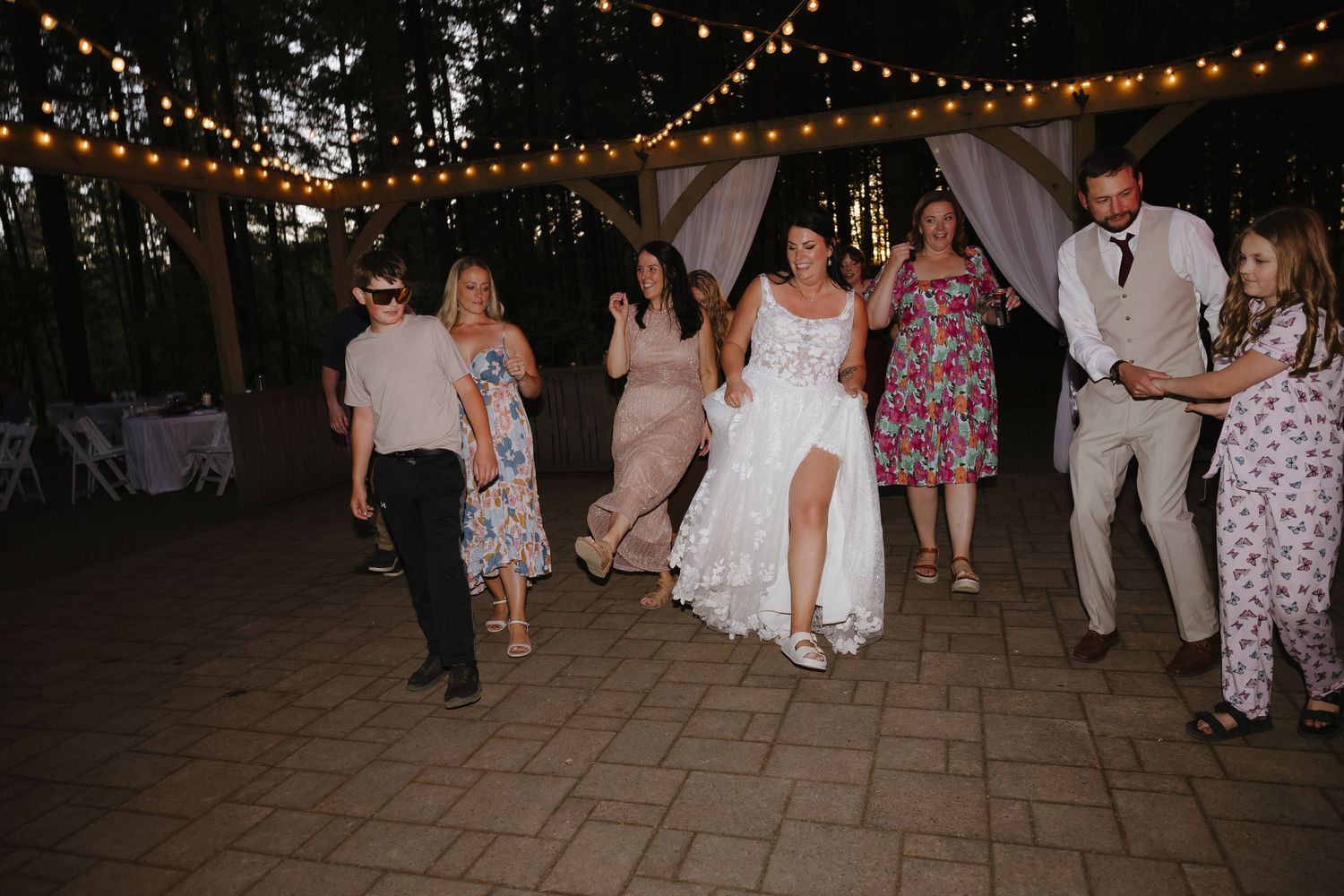 Bride lifts her lace gown while dancing with guests under string lights at an outdoor nighttime forest reception.