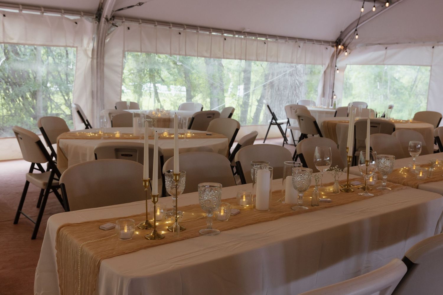 Elegant wedding reception tent interior with round tables, white chairs, candles, and string lights along the ceiling.