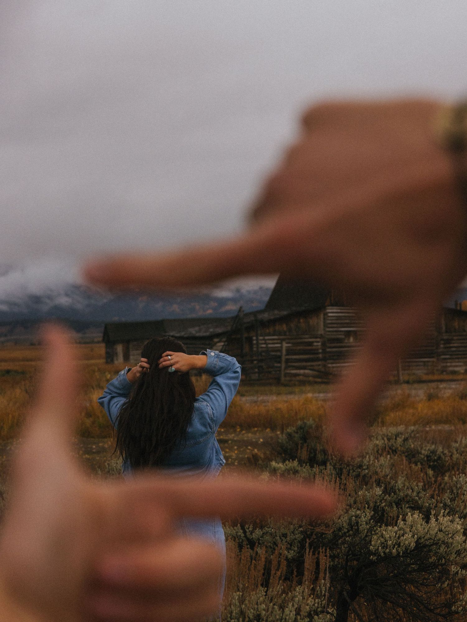 Creative framing shot of woman adjusting hair in field with rustic barn, hands forming a frame foreground.