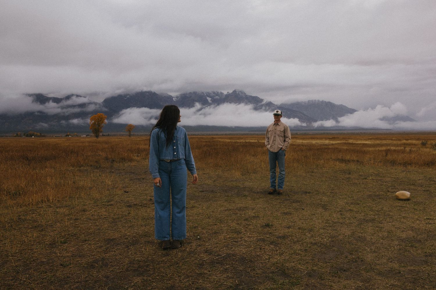 Couple standing apart in a vast golden meadow with dramatic cloud-covered mountains behind them.