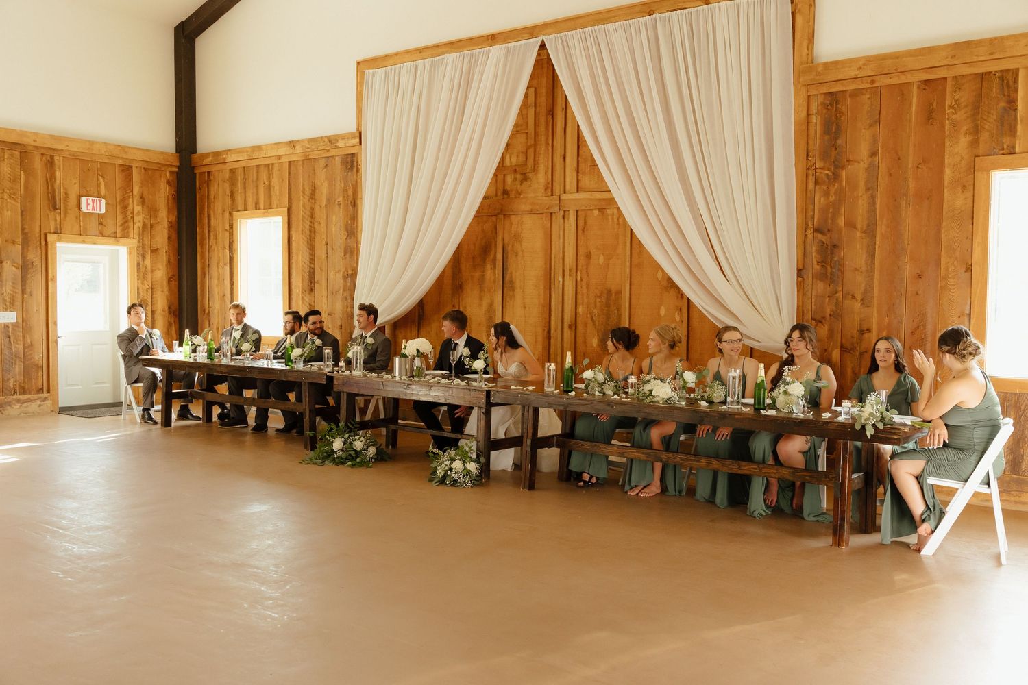 Wedding party seated at long head table in wood-paneled barn with elegant draping backdrop.